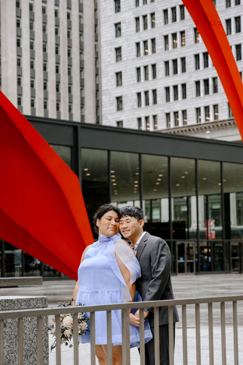A couple poses together beneath a striking red sculpture against an urban backdrop.