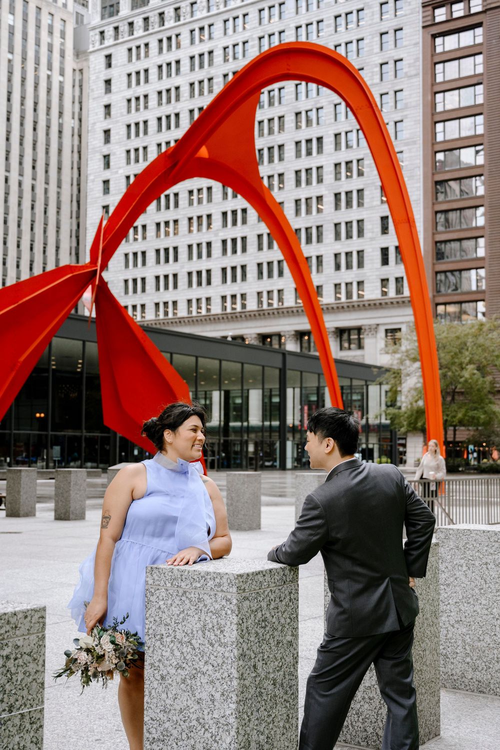 A couple poses by concrete barriers under dramatic red sculptural arches with skyscrapers in the background.