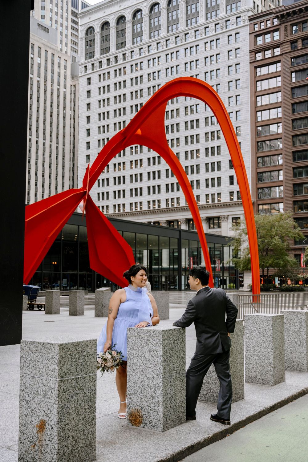 A couple stands by concrete blocks under a striking red metal sculpture with city buildings in the background.