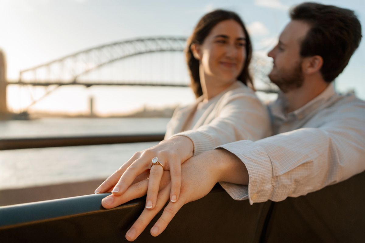 proposal photographer in Sydney photo of a engagement ring in focus while couple blurry in Sydney with the harbour bridge in the background
