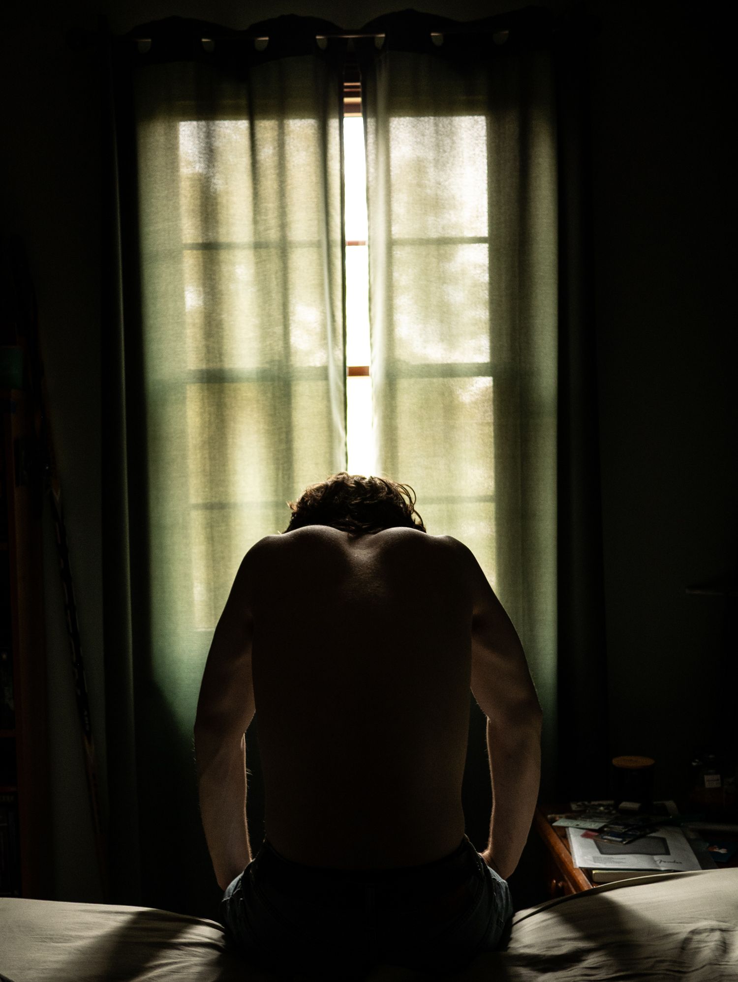 Shirtless man in the dark lit by a window with closed curtains