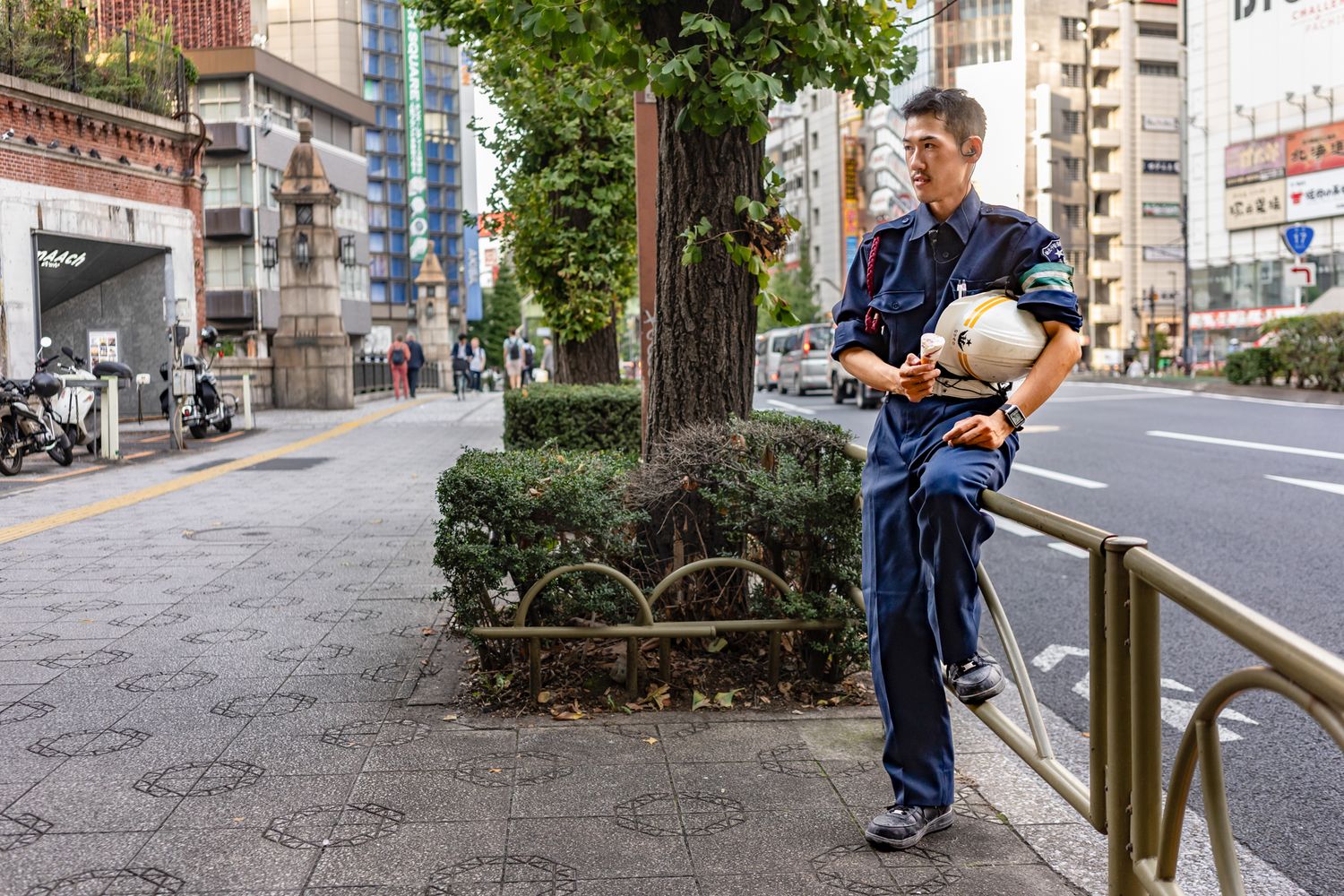 Professional delivery person in blue uniform stands on urban street with buildings and trees in background.