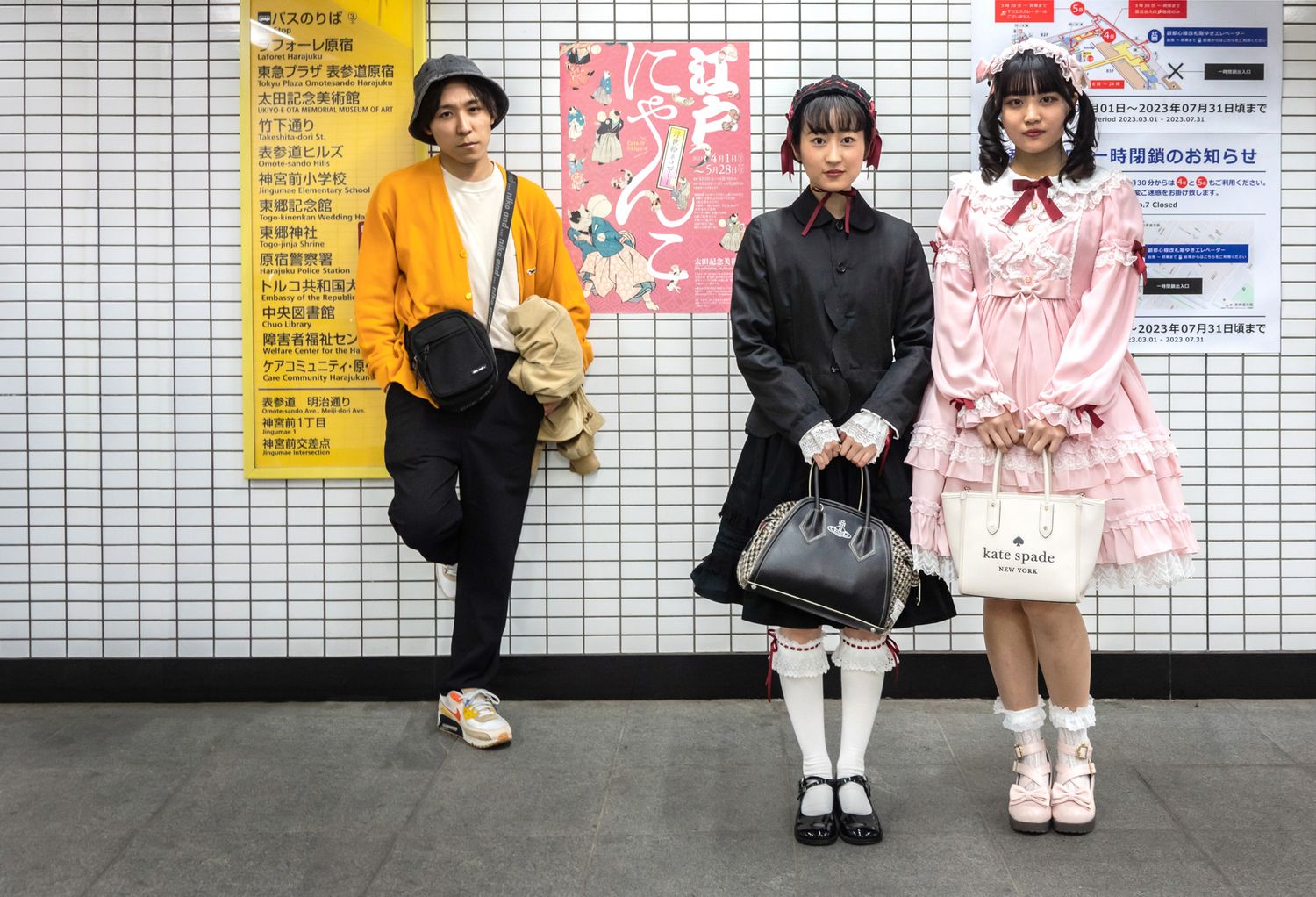 Three fashionably dressed people posing against a tiled wall with posters in a subway station.
