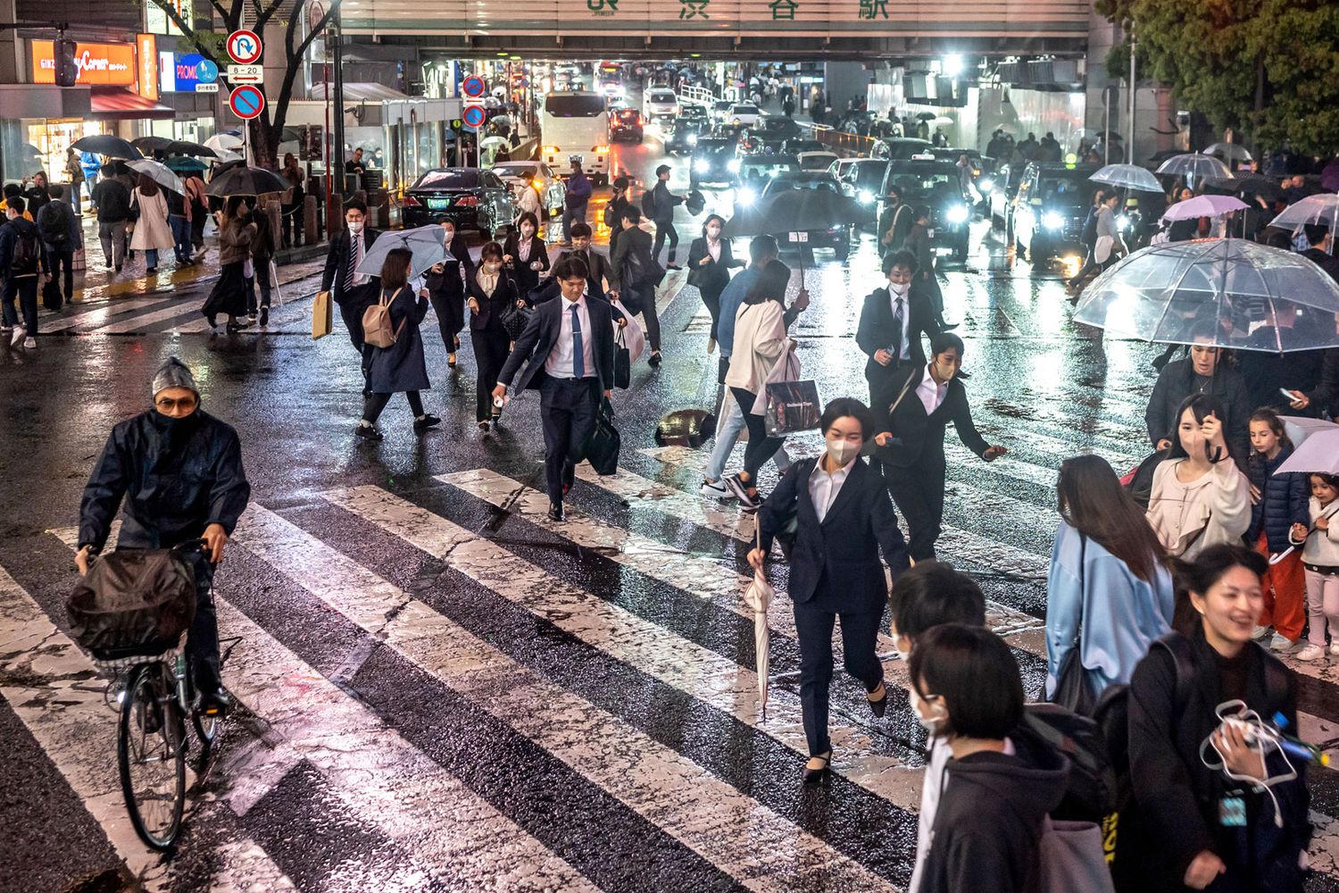 Rainy night street scene with crowd crossing illuminated crosswalk under umbrellas.