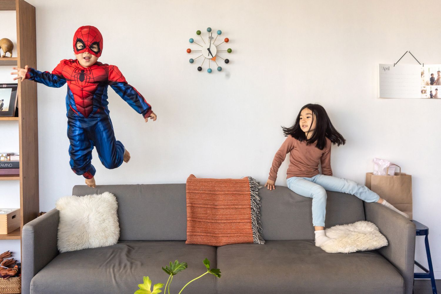 Children jumping on gray couch in living room with one wearing superhero costume.