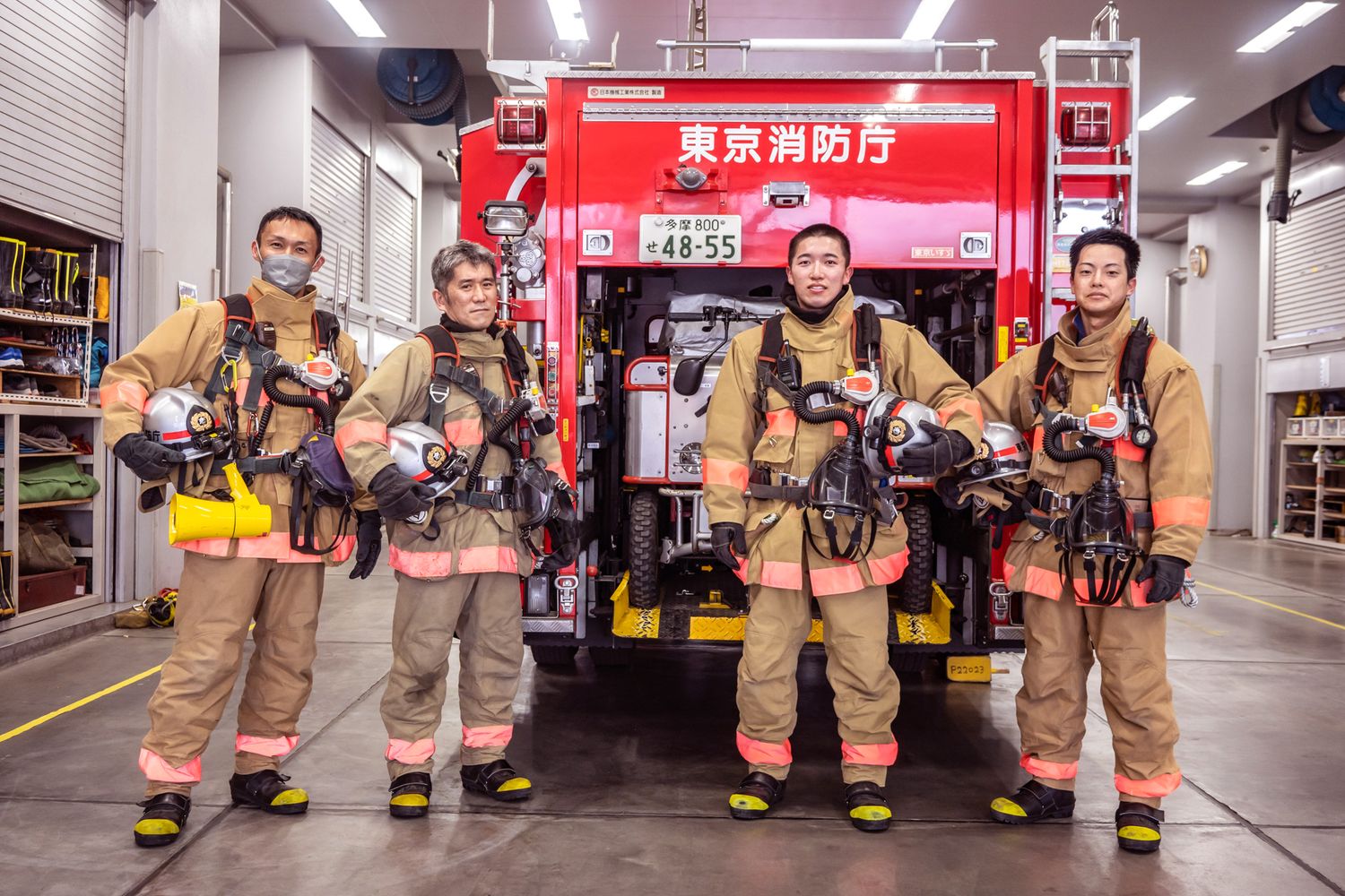 Firefighters pose with their equipment in front of a red fire truck in a station bay.