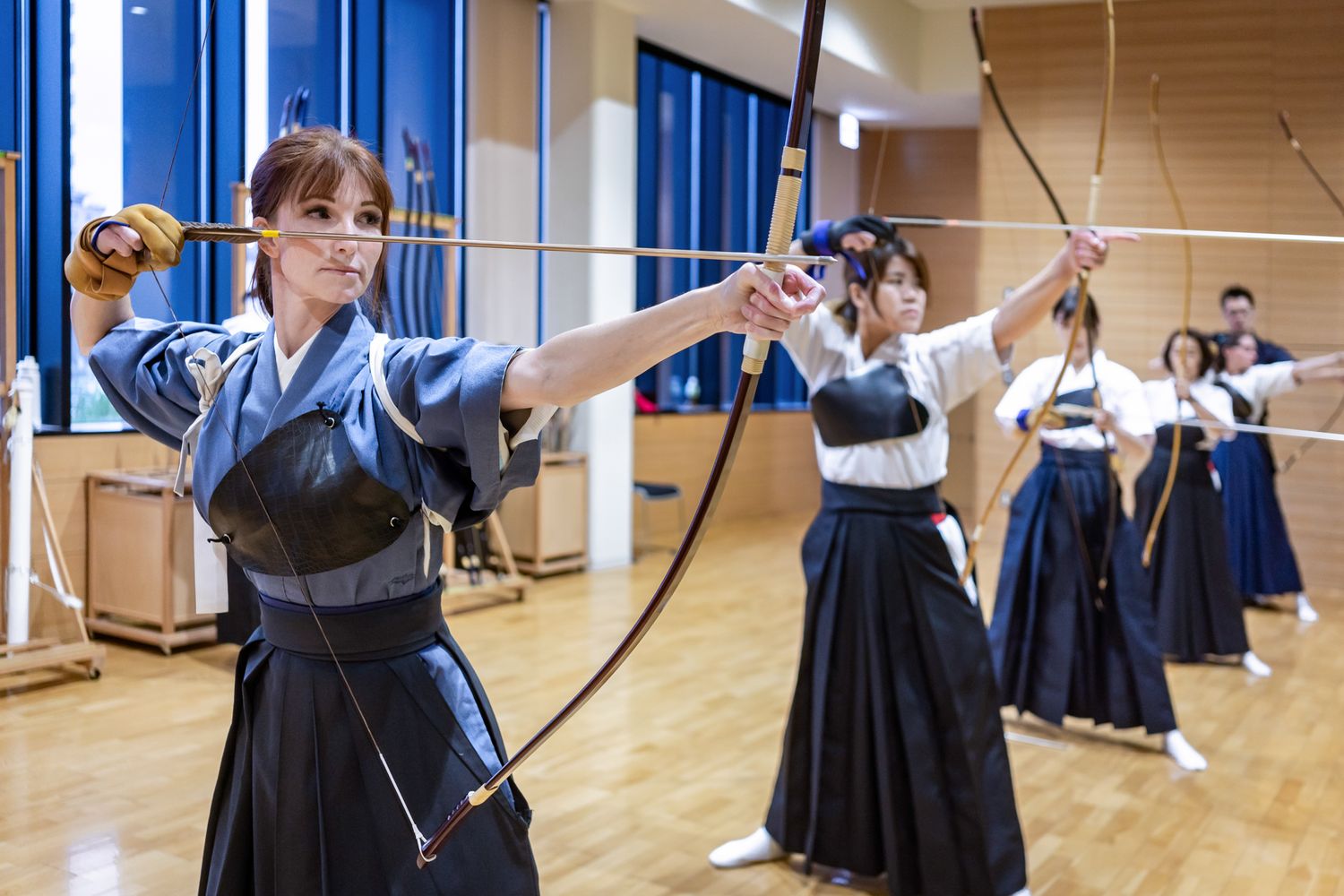 Students practicing kyudo archery in indoor training facility wearing traditional hakama.