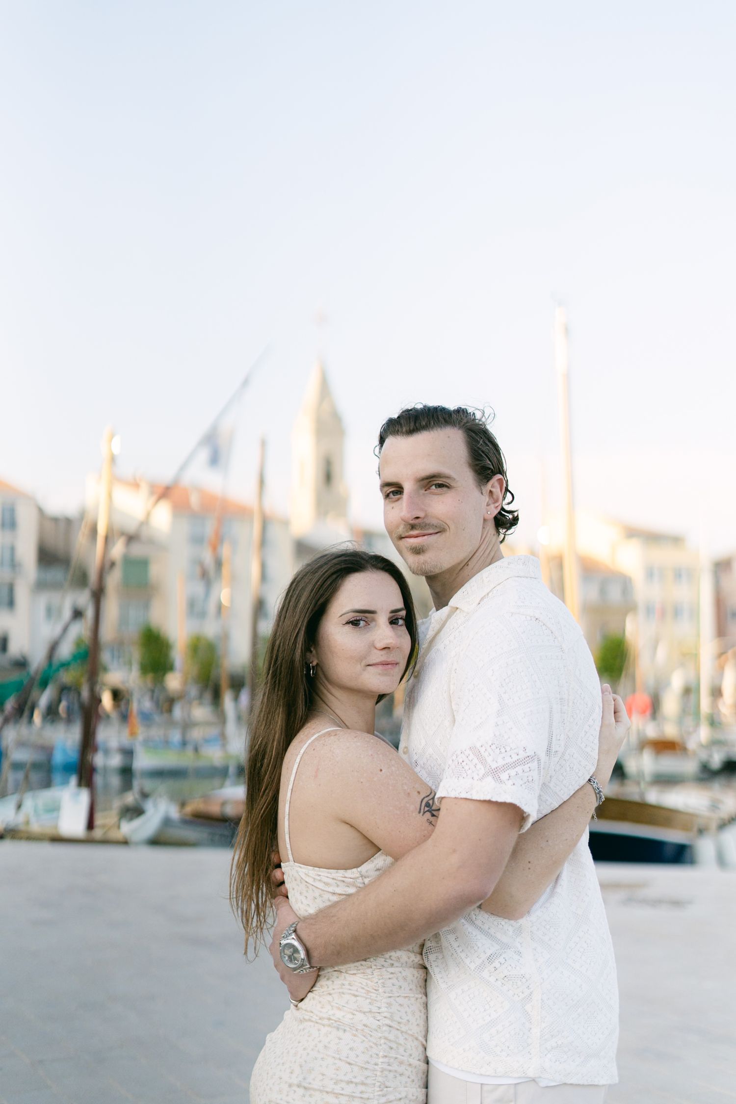 A romantic couple embraces at a marina with boats and buildings in the background at sunset.