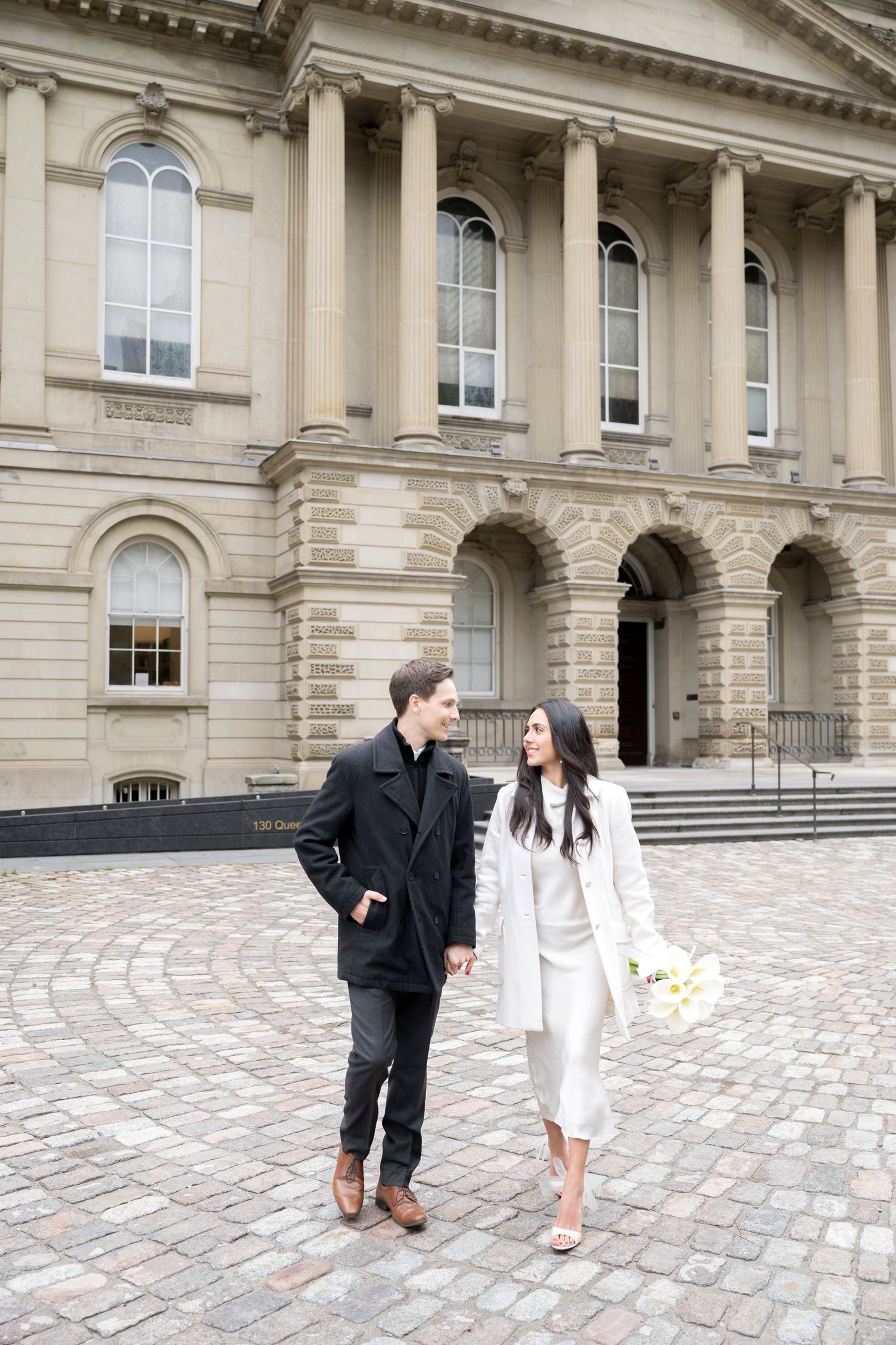 Couple in formal attire walking together on cobblestone courtyard of historic building with neoclassical architecture.