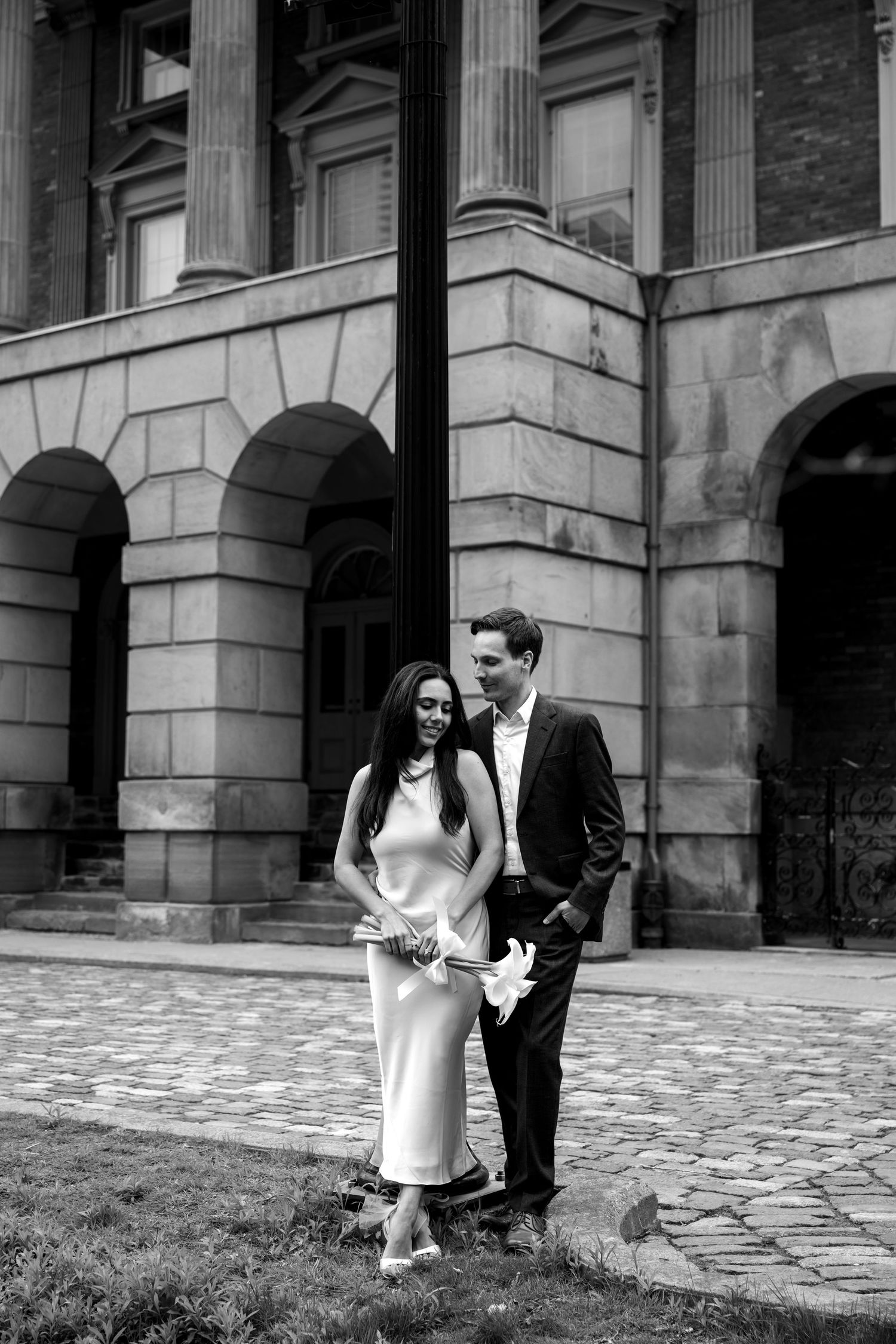 A couple in formal attire stands together outside a historic stone building with arched columns and cobblestone path.