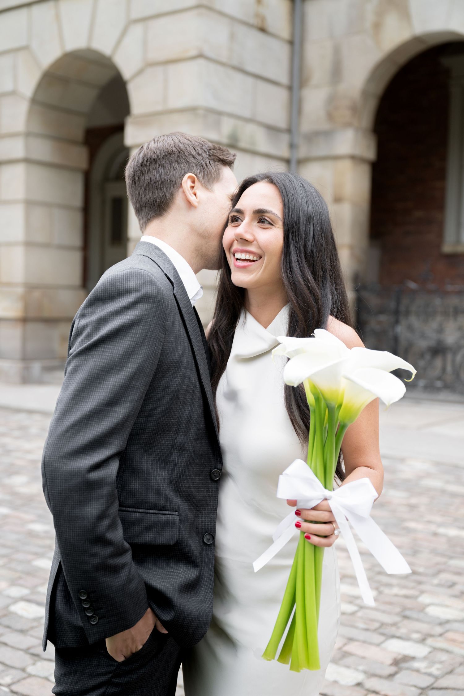 Happy couple sharing intimate moment while holding calla lily bouquet in front of stone building.