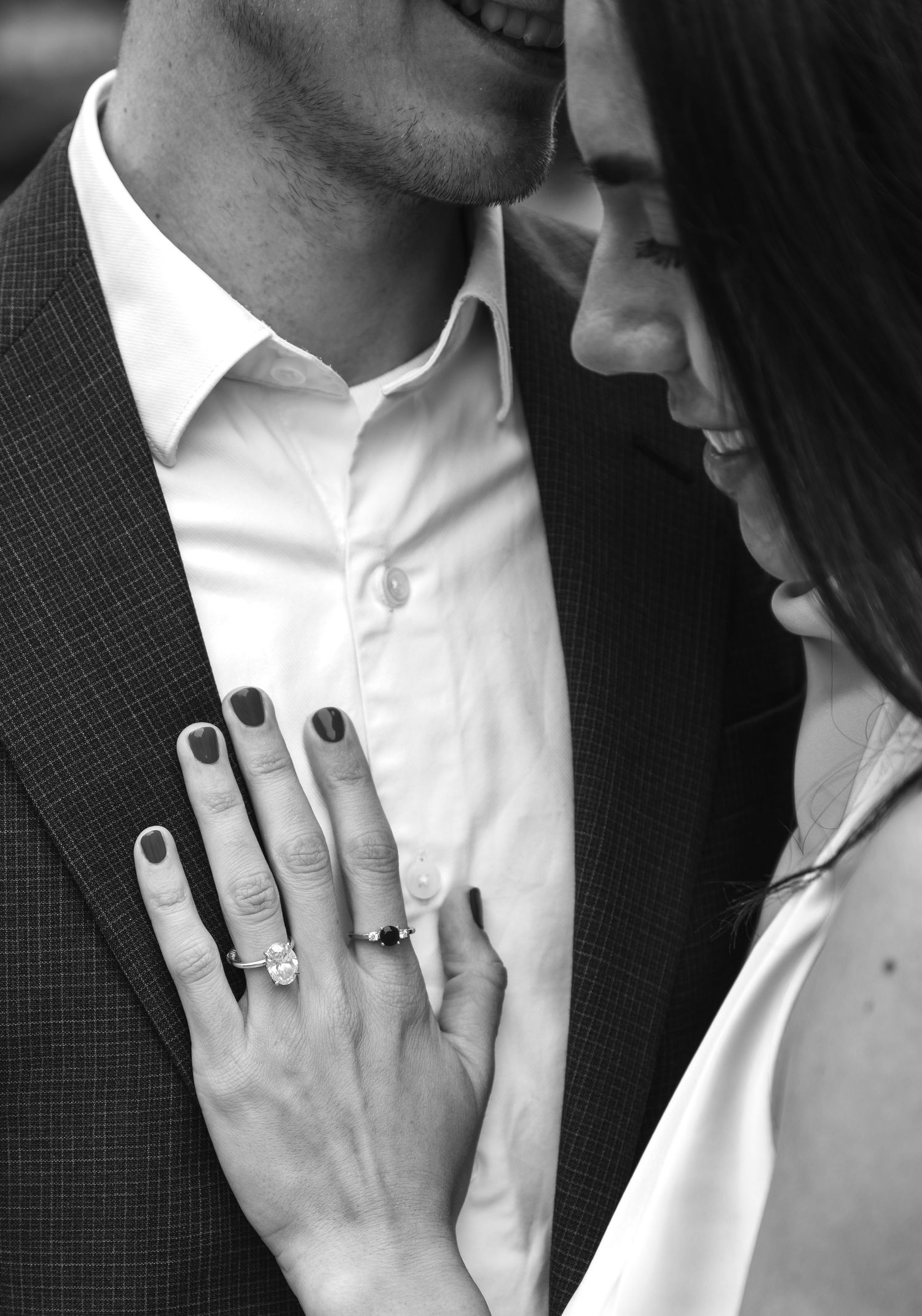 A black and white close-up shot of an engaged couple showing her diamond ring against his suit jacket.