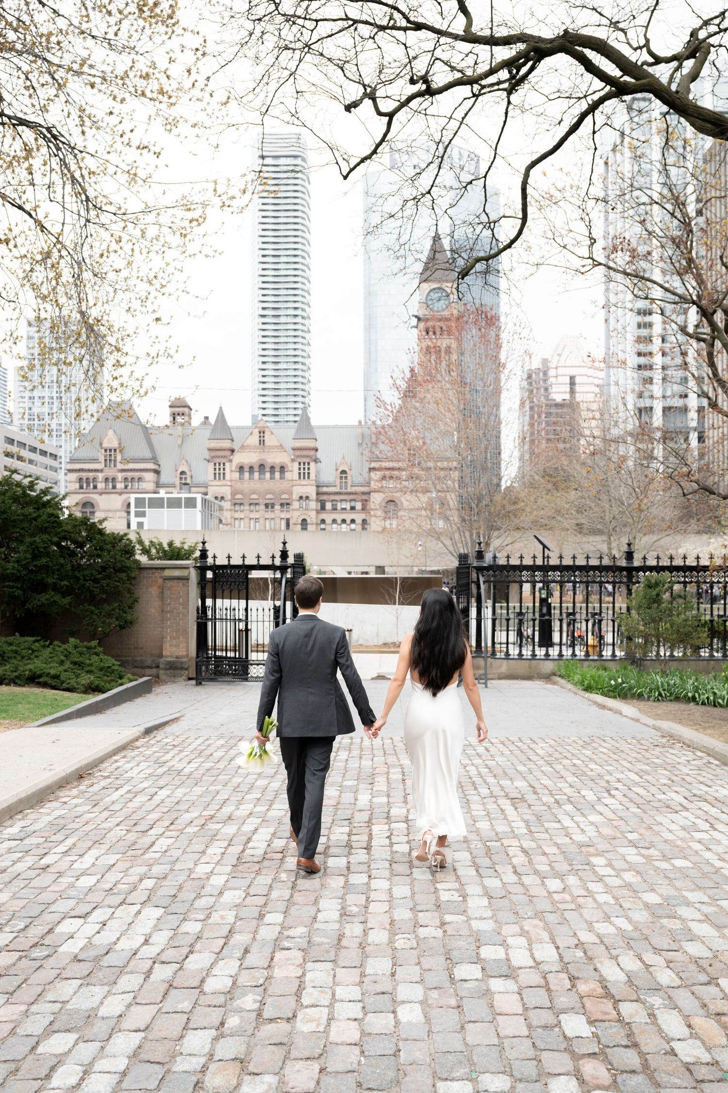 A couple walks hand in hand down a cobblestone path with NYC skyscrapers and architecture in the background.