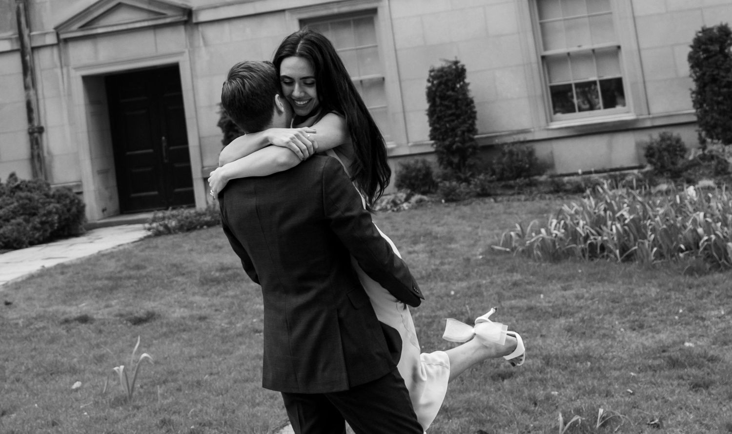 A joyful couple embraces in black and white on a lawn in front of a building with the person being lifted off their feet.