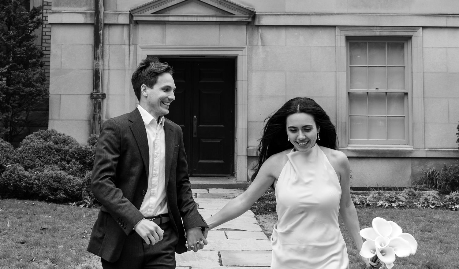 A black and white photograph of a couple running joyfully in front of a building entrance.