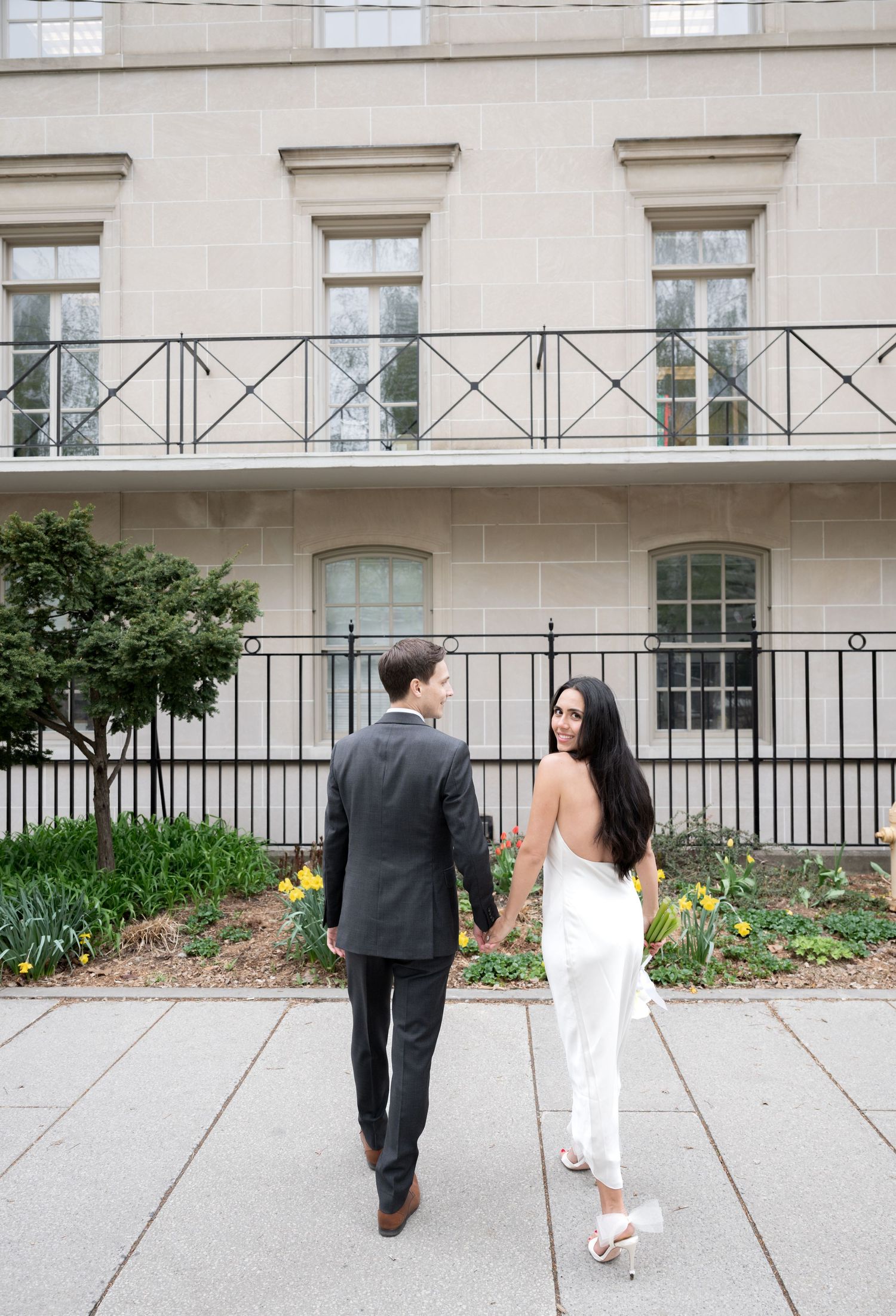 A couple walks together along a sidewalk next to a historic building with elegant architecture and landscaping.