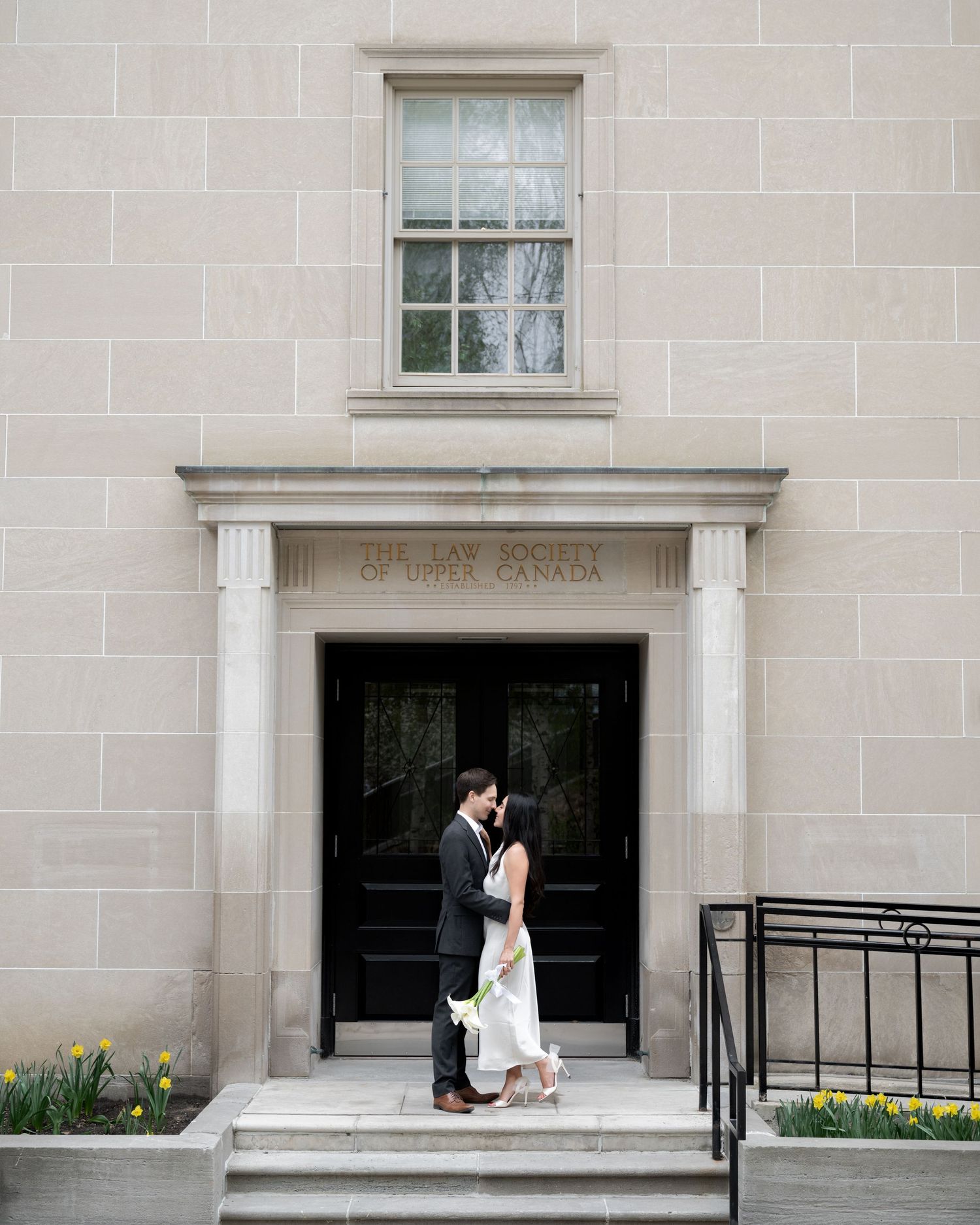 A couple stands in a romantic pose under a classical stone doorway entrance with columns.