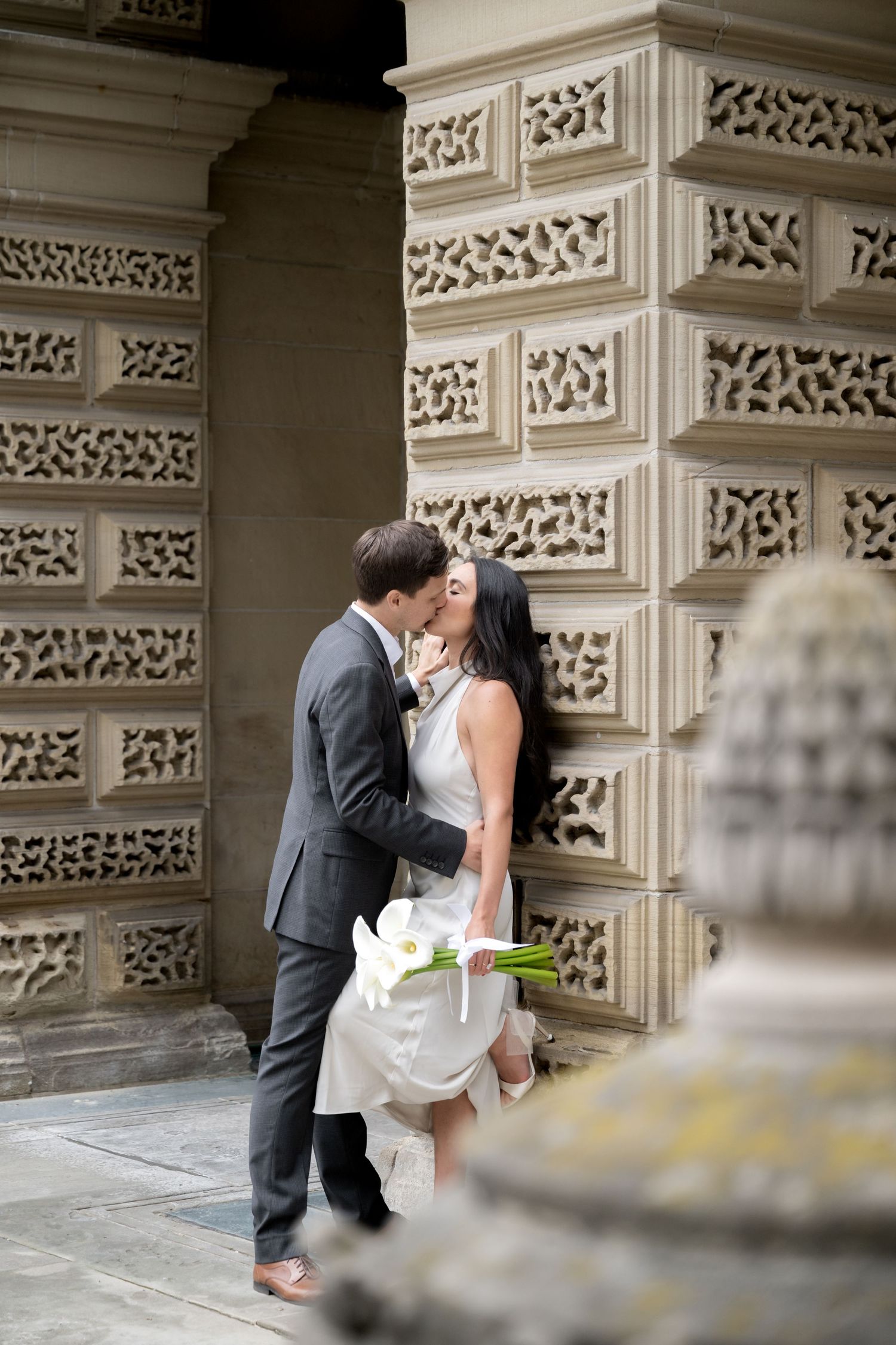 Newlyweds share an intimate kiss against an ornate stone wall with decorative carved patterns while bride holds white bouquet.