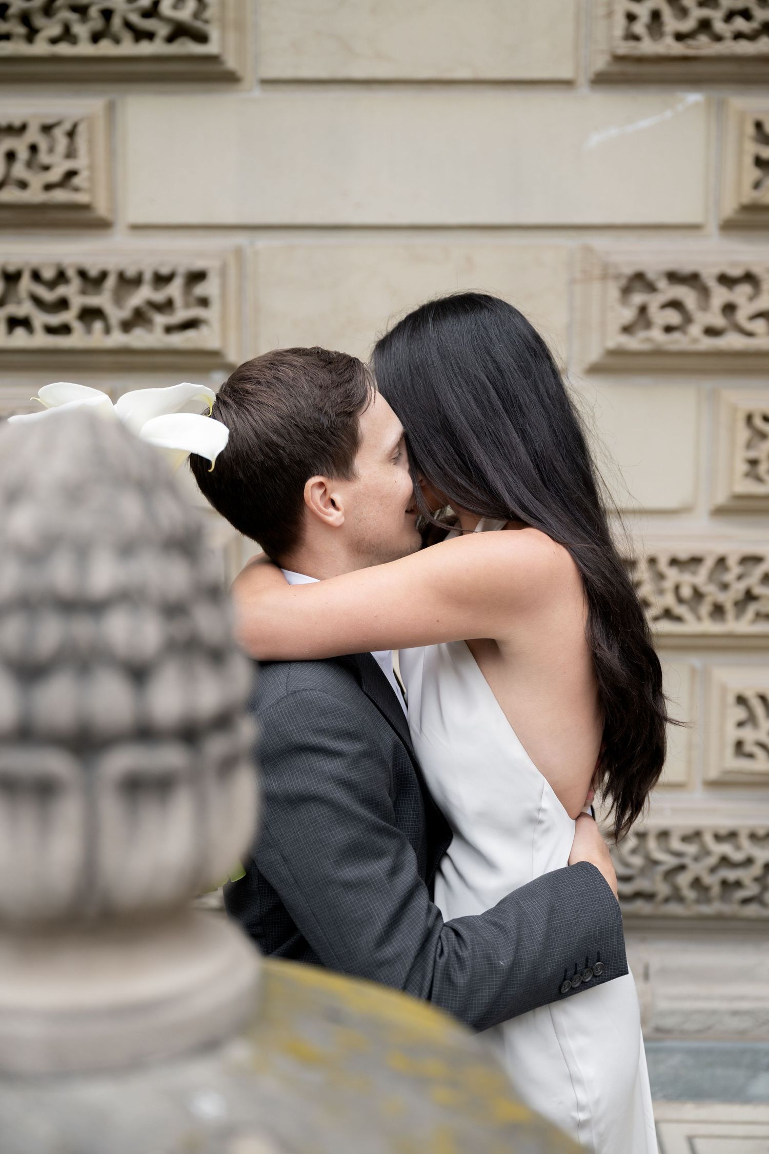 Two people share an intimate embrace while kissing in front of an ornate stone building facade.