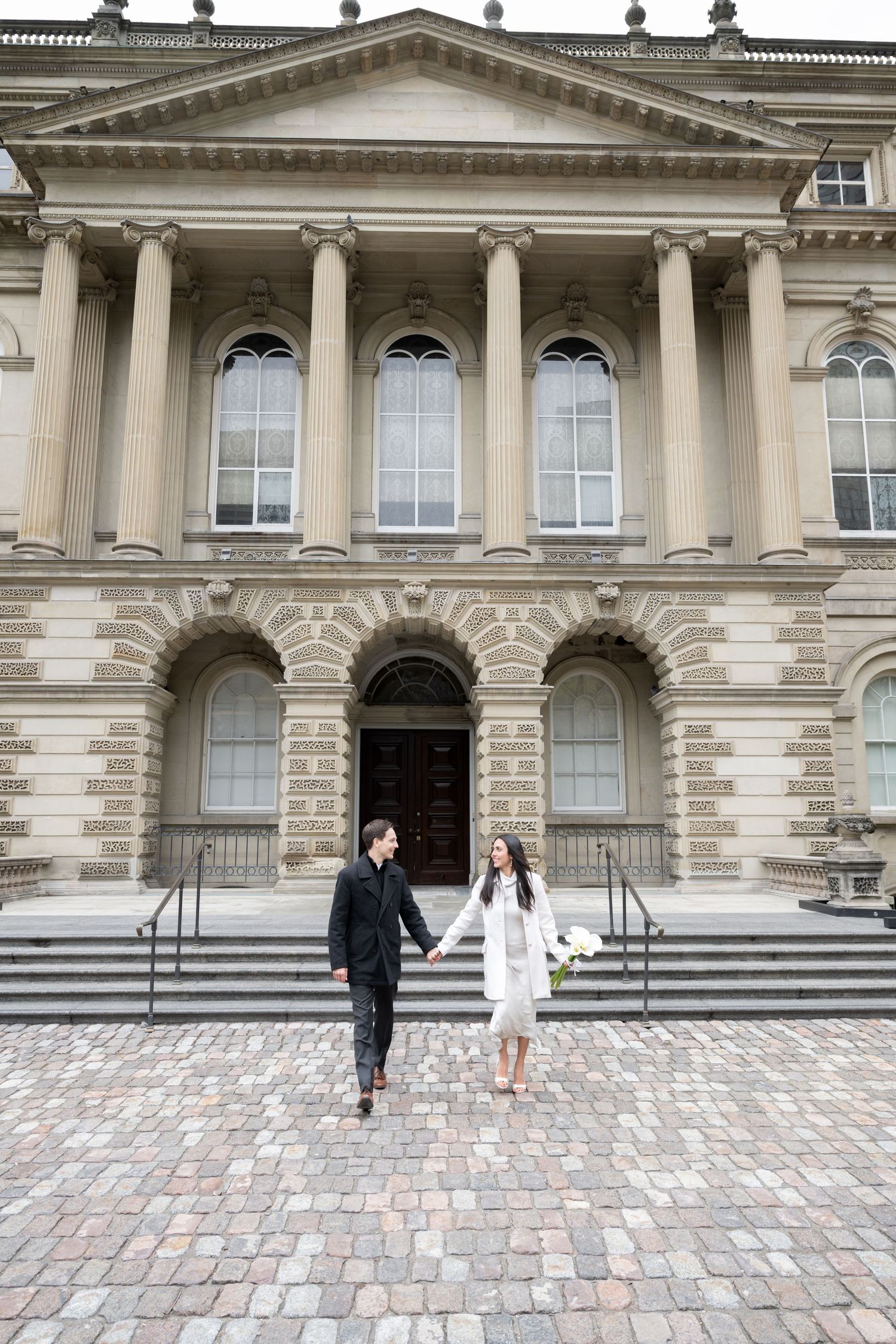 A couple in formal attire stands on a brick courtyard in front of a grand neoclassical building with tall columns.