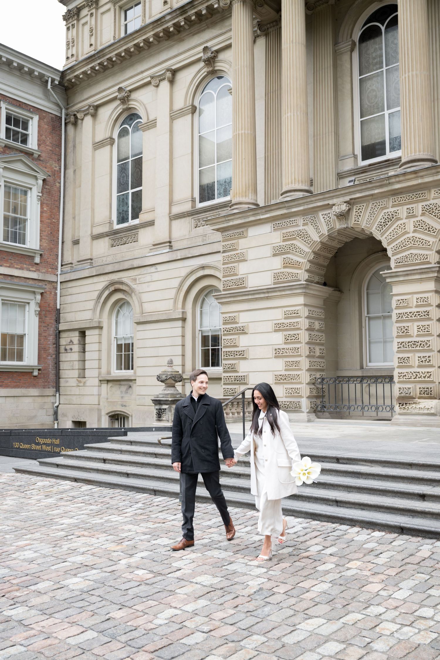 Two people walk down stone steps of a grand neoclassical building with tall arched windows and columns.