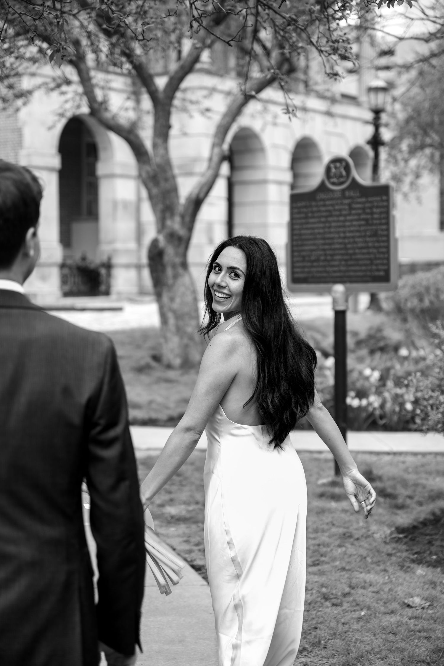 Black and white wedding photo showing a bride in a white dress looking back over her shoulder in front of an arched building.