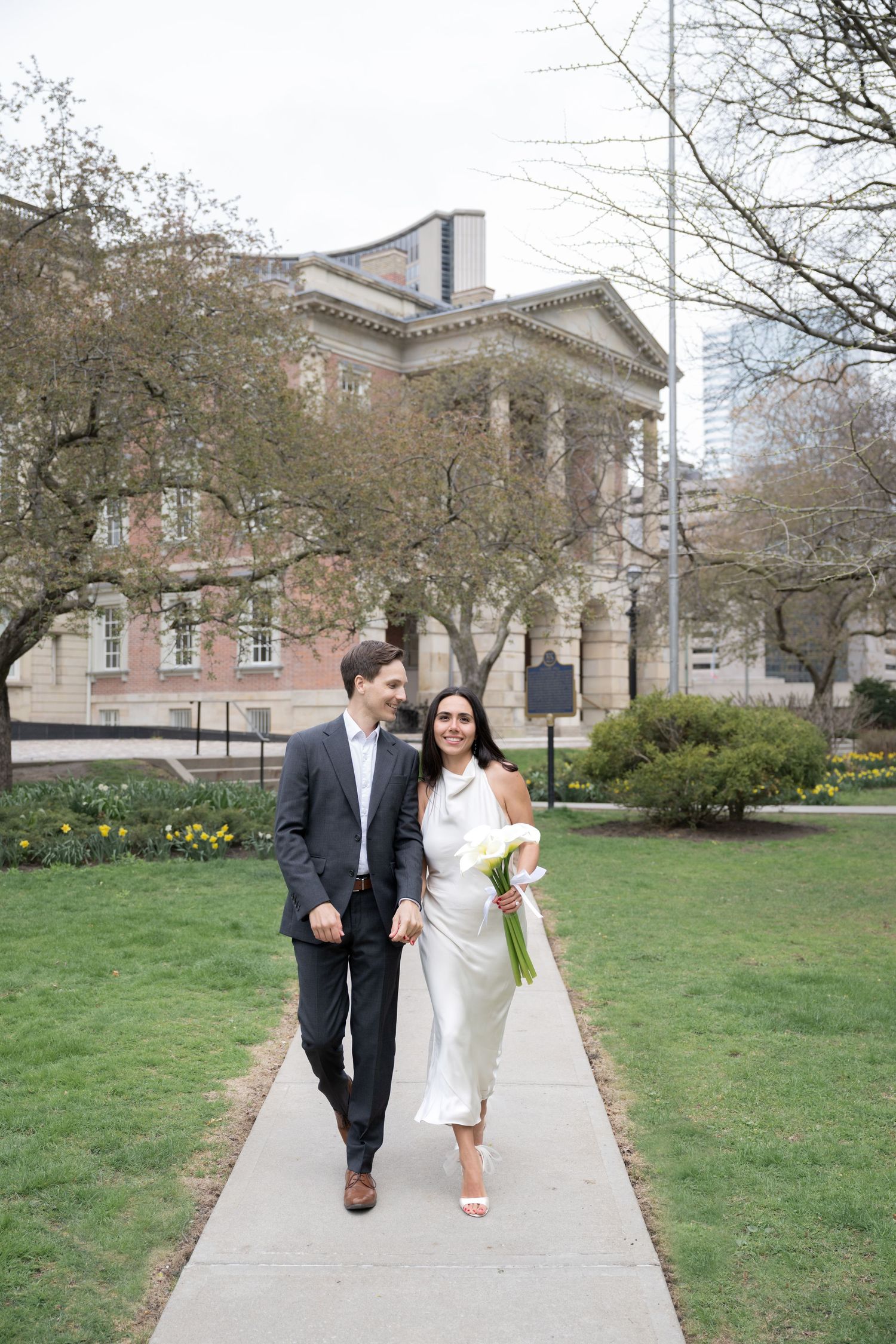 A couple walks along a pathway in front of a grand neoclassical building with columns and spring foliage.