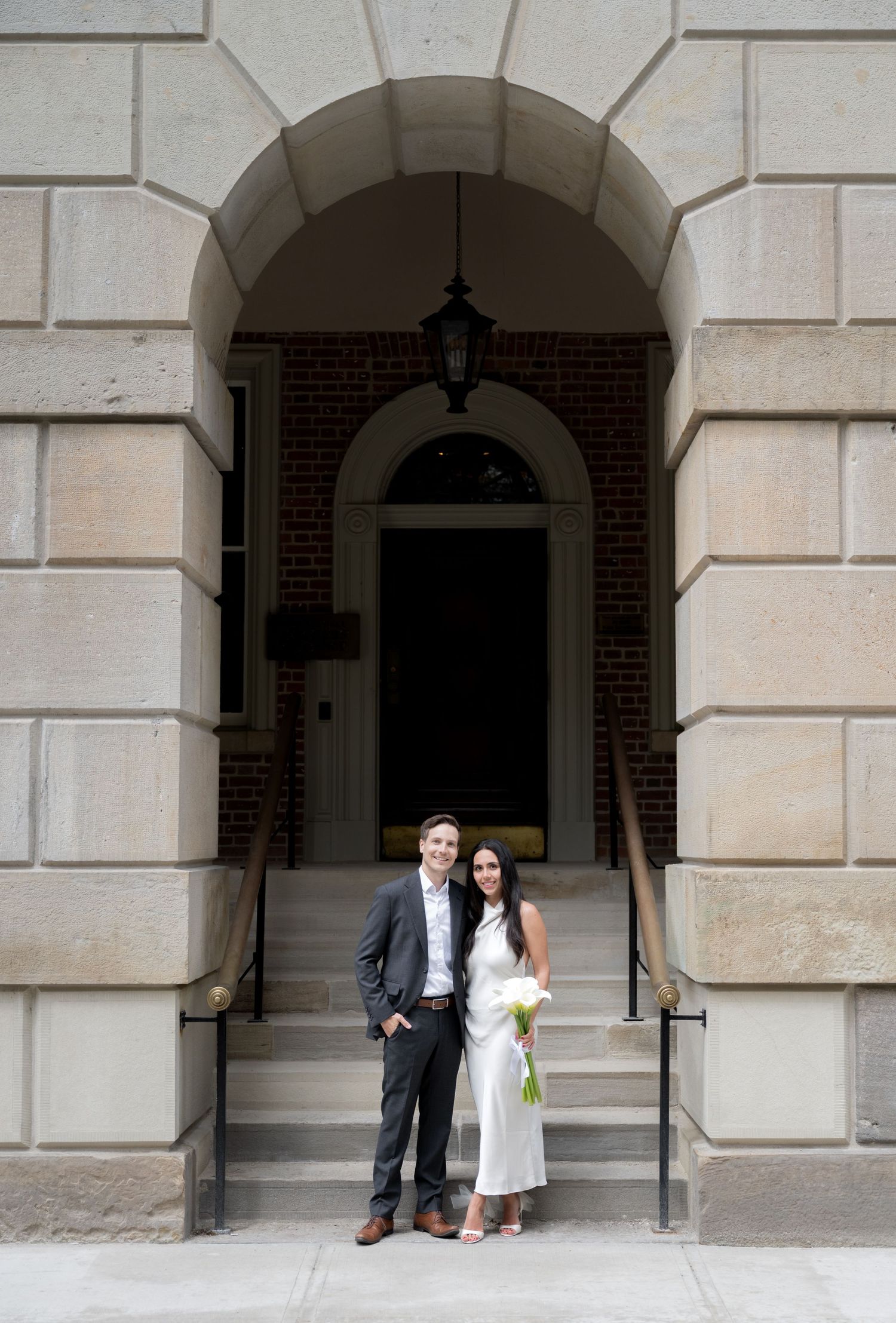 A couple stands together under an elegant archway entrance with white stone columns and brick detailing.