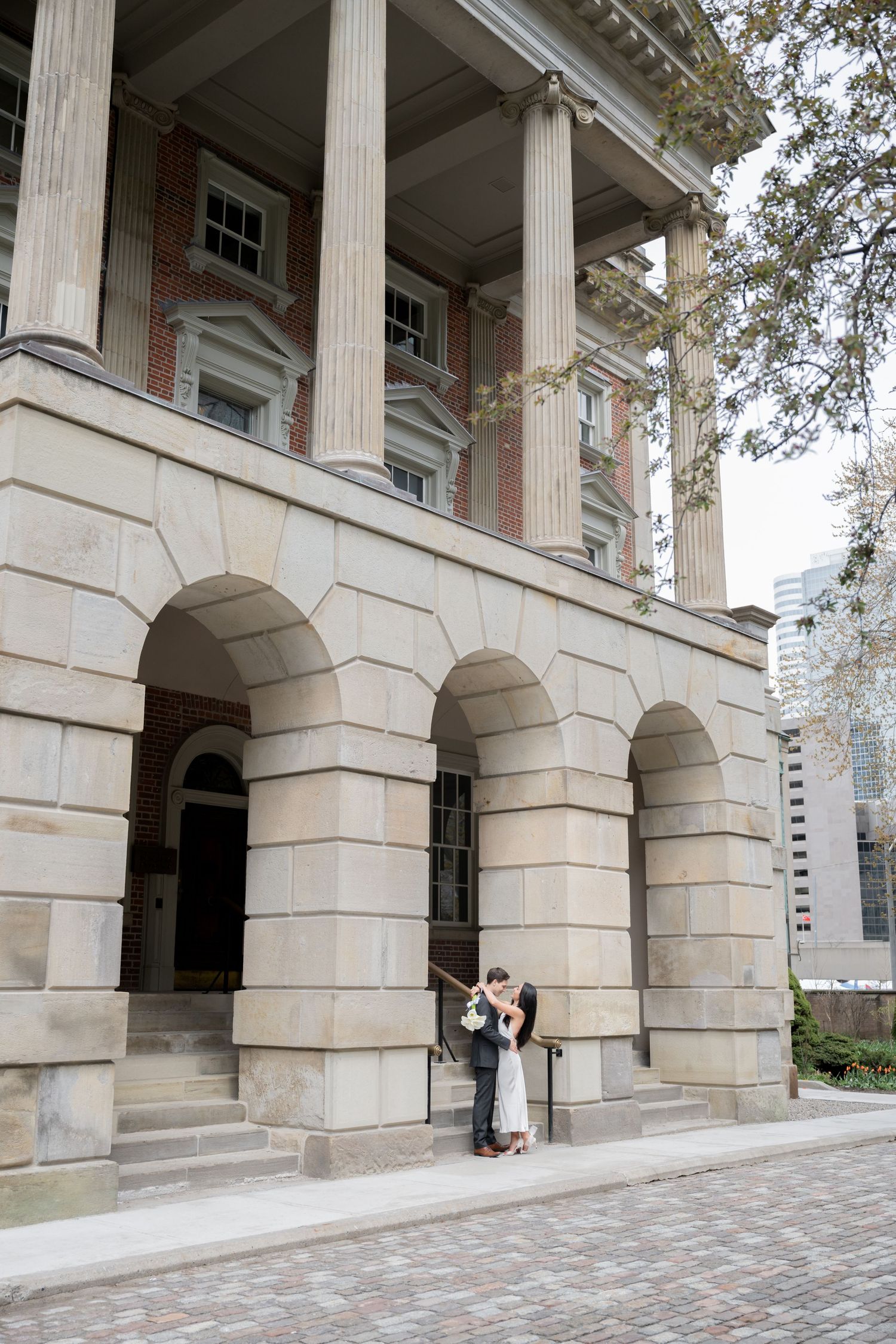 A classical building exterior with grand columns and stone arches frames two people standing beneath its elegant architecture.