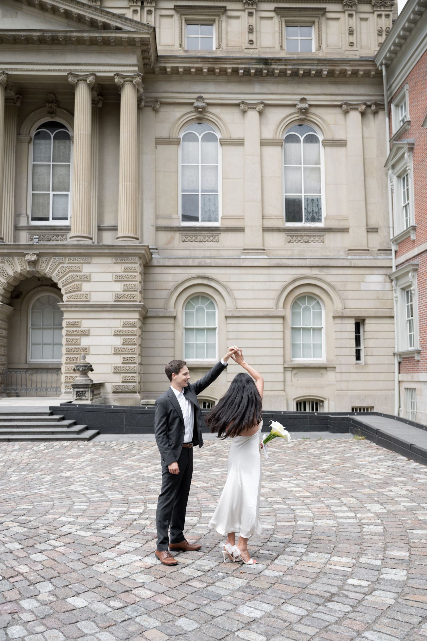 A couple dances on a cobblestone courtyard in front of a historic building with arched windows and ornate stonework.