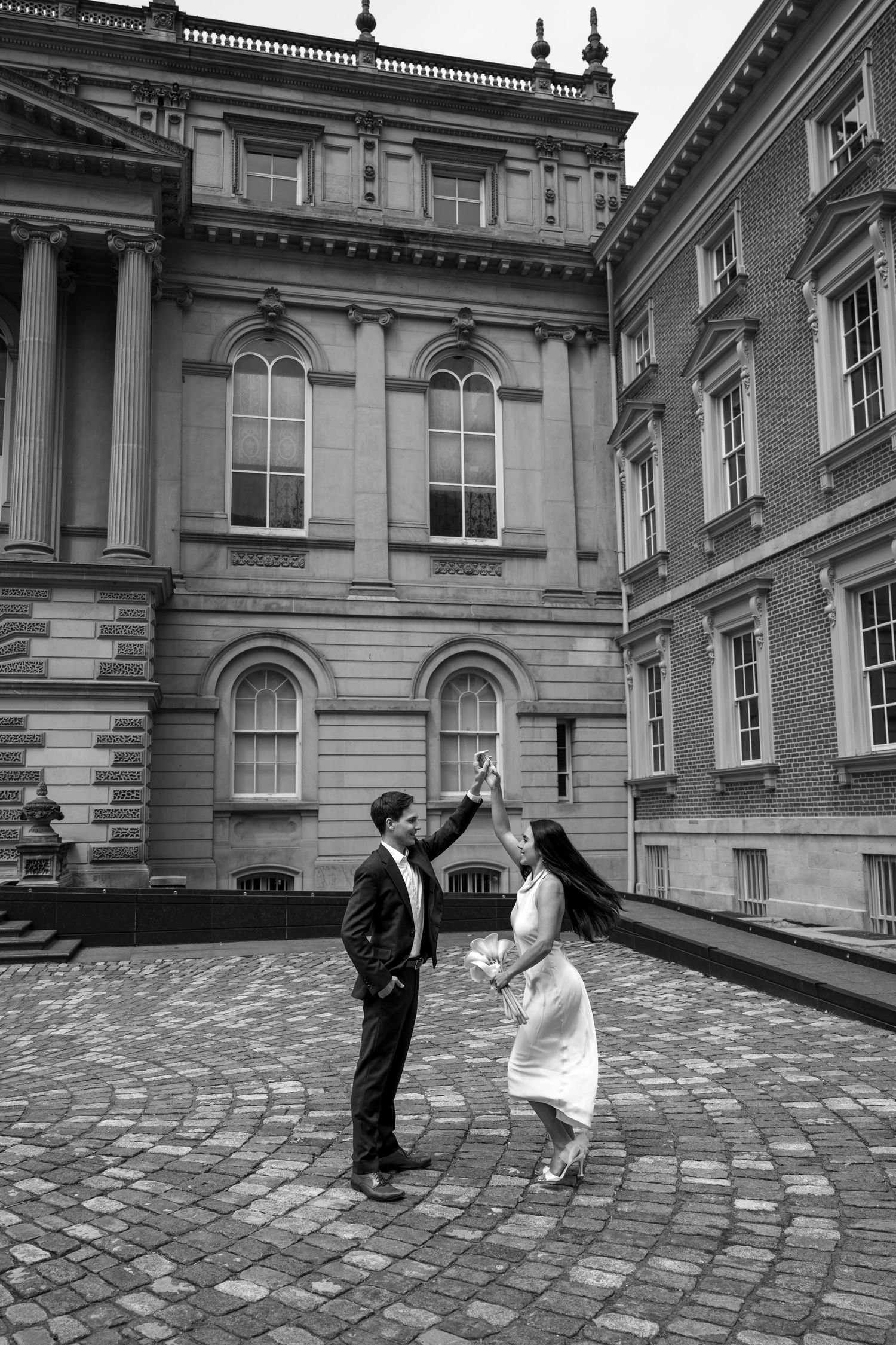 Playful couple dancing together on cobblestone courtyard in front of historic building in black and white.