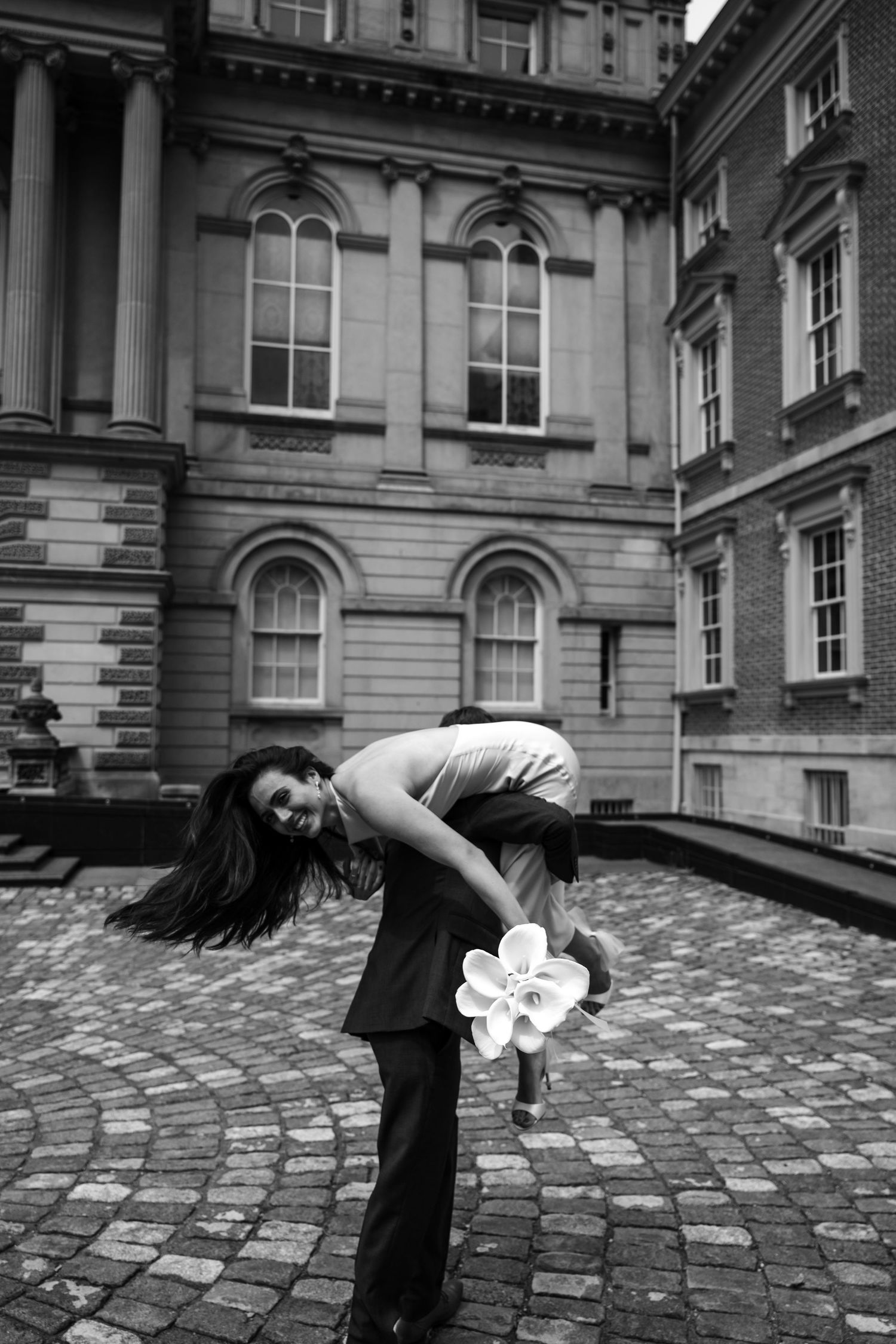 A graceful figure in dark attire bends forward holding flowers against the backdrop of a historic stone building exterior.