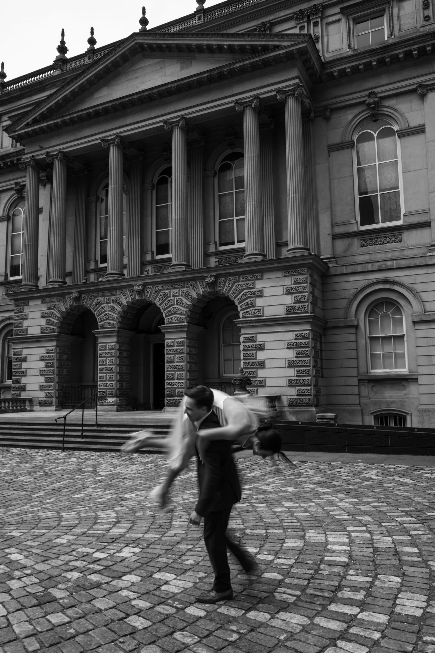 A person spins joyfully on cobblestones in front of a grand neoclassical building with tall columns and arched windows.