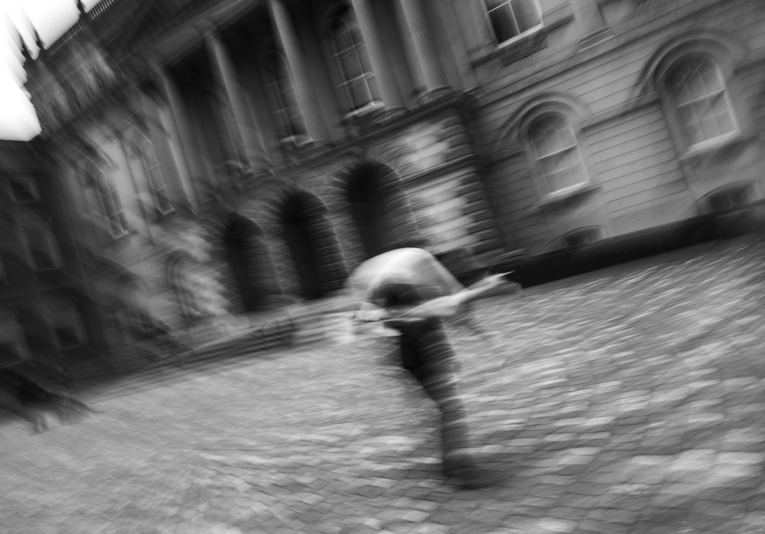 A blurred black and white motion shot shows a person with an umbrella walking on a cobblestone street past historic buildings.