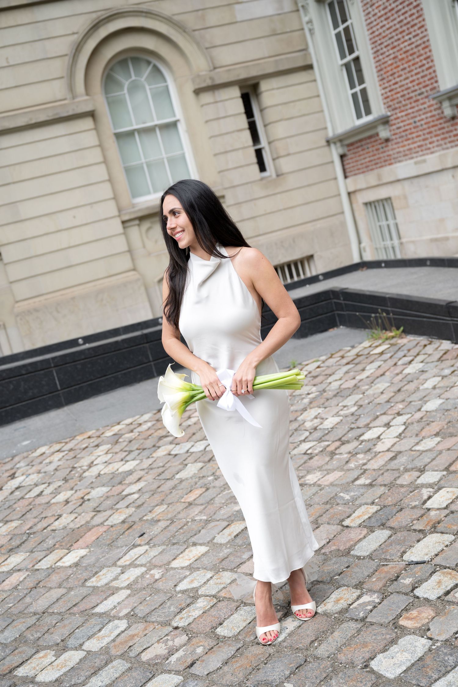Woman in elegant white dress holding calla lilies poses on cobblestone courtyard near historic building.