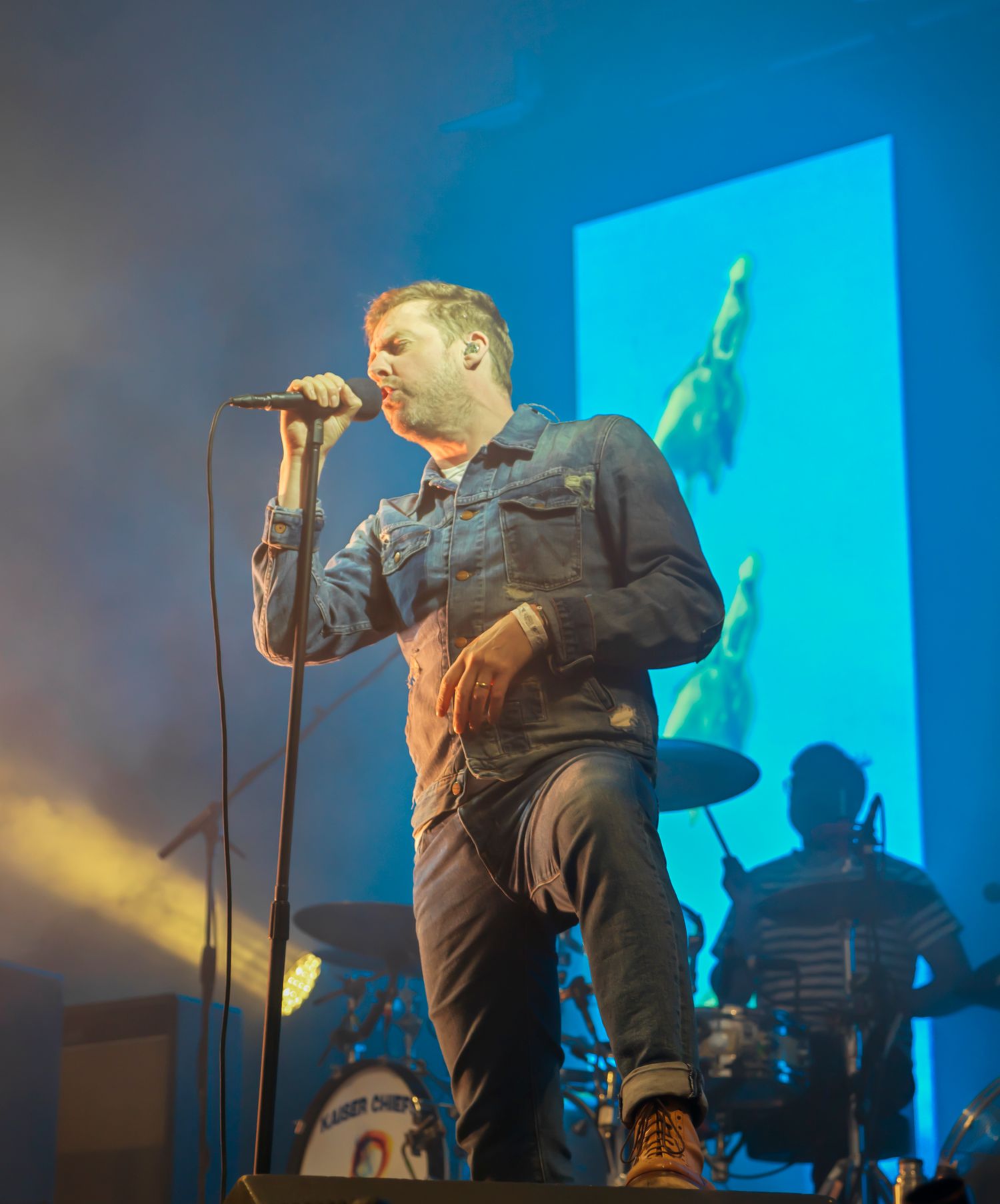 Lead singer performing on stage with blue projection screen and band silhouettes in background.