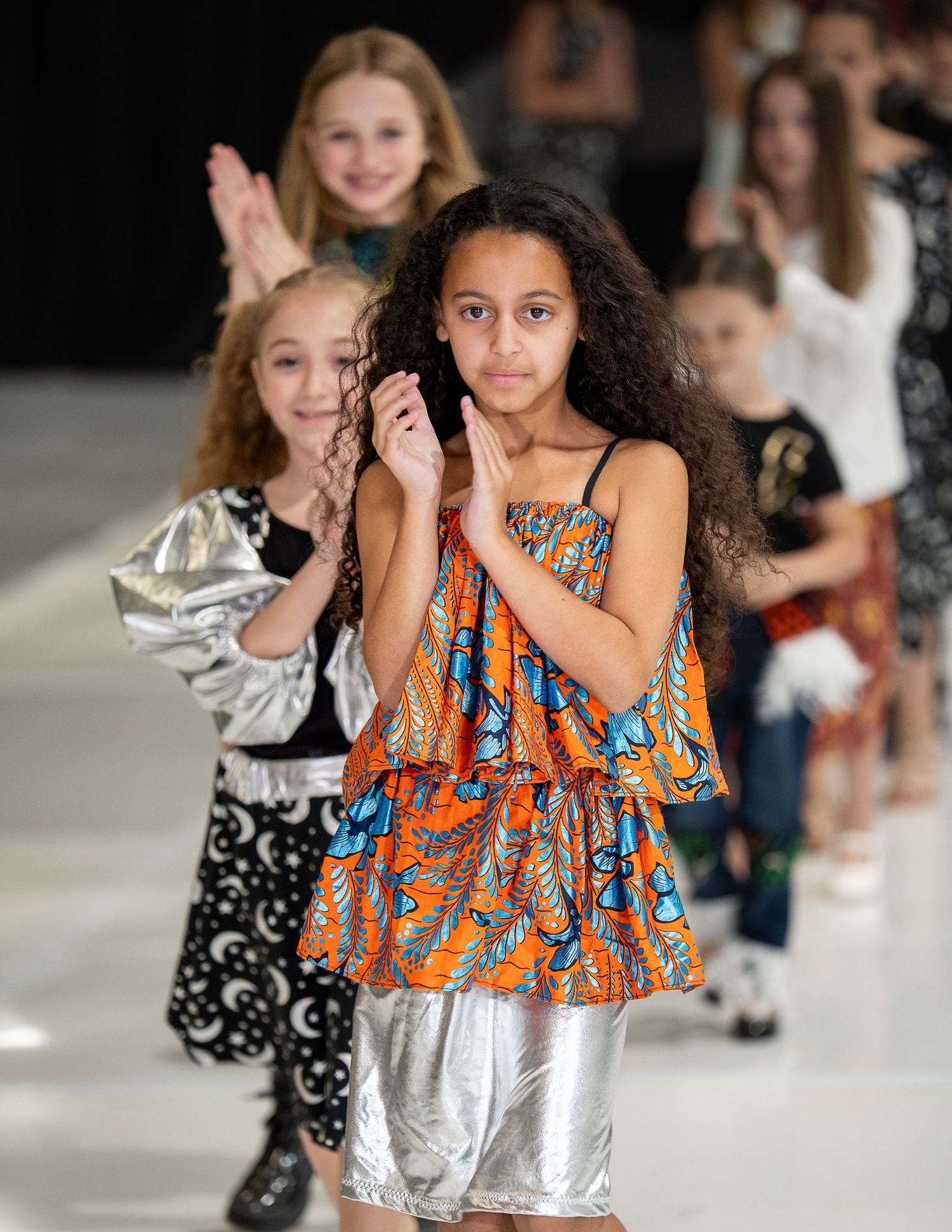 Models walk down a fashion runway wearing colorful designs including an orange patterned dress and metallic pants.