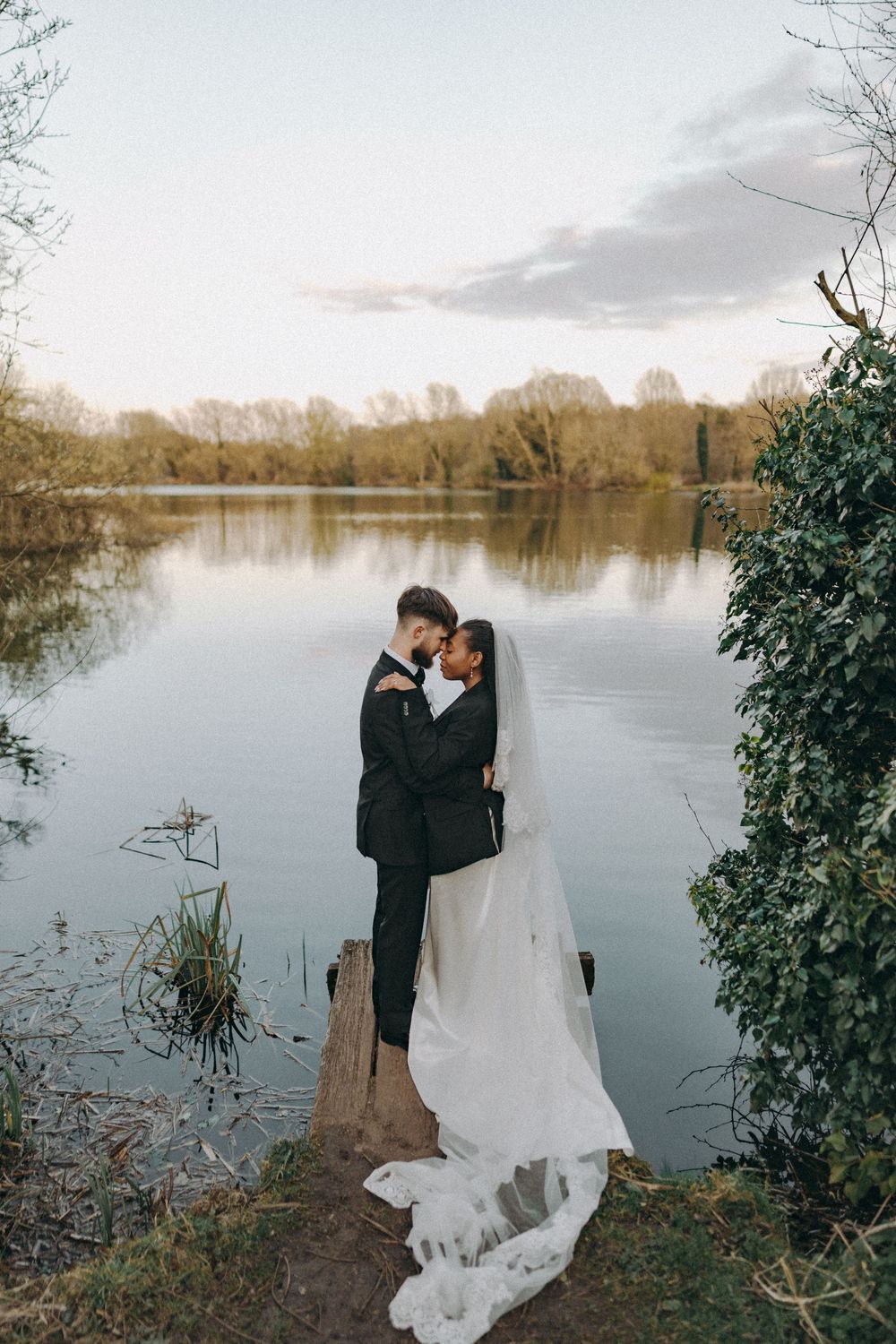 Wide photo of Bride groom in an embrace, foreheads adjoined to the backdrop of a lake. Documentary wedding