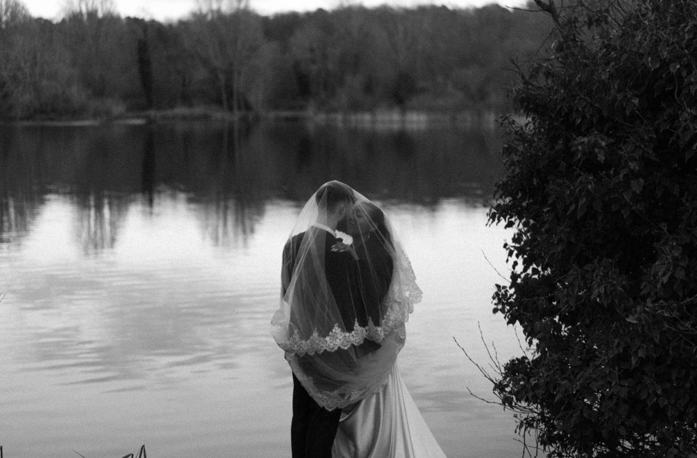 B&W photo of bride groom sharing kiss (under veil) to the backdrop of a lake. Documentary wedding