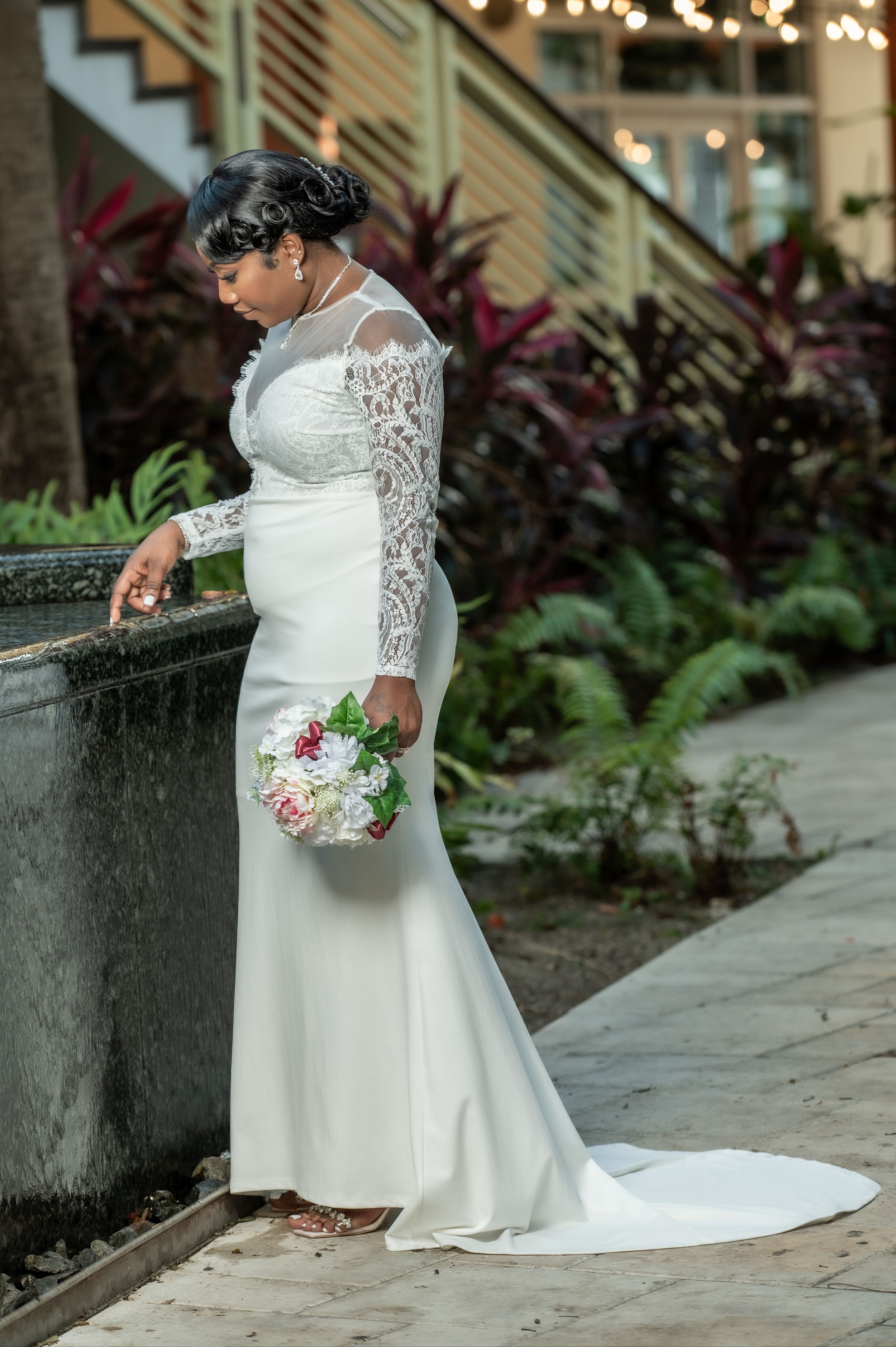 Bride in long-sleeve lace wedding gown with train holds bouquet outdoors near water feature with tropical greenery.
