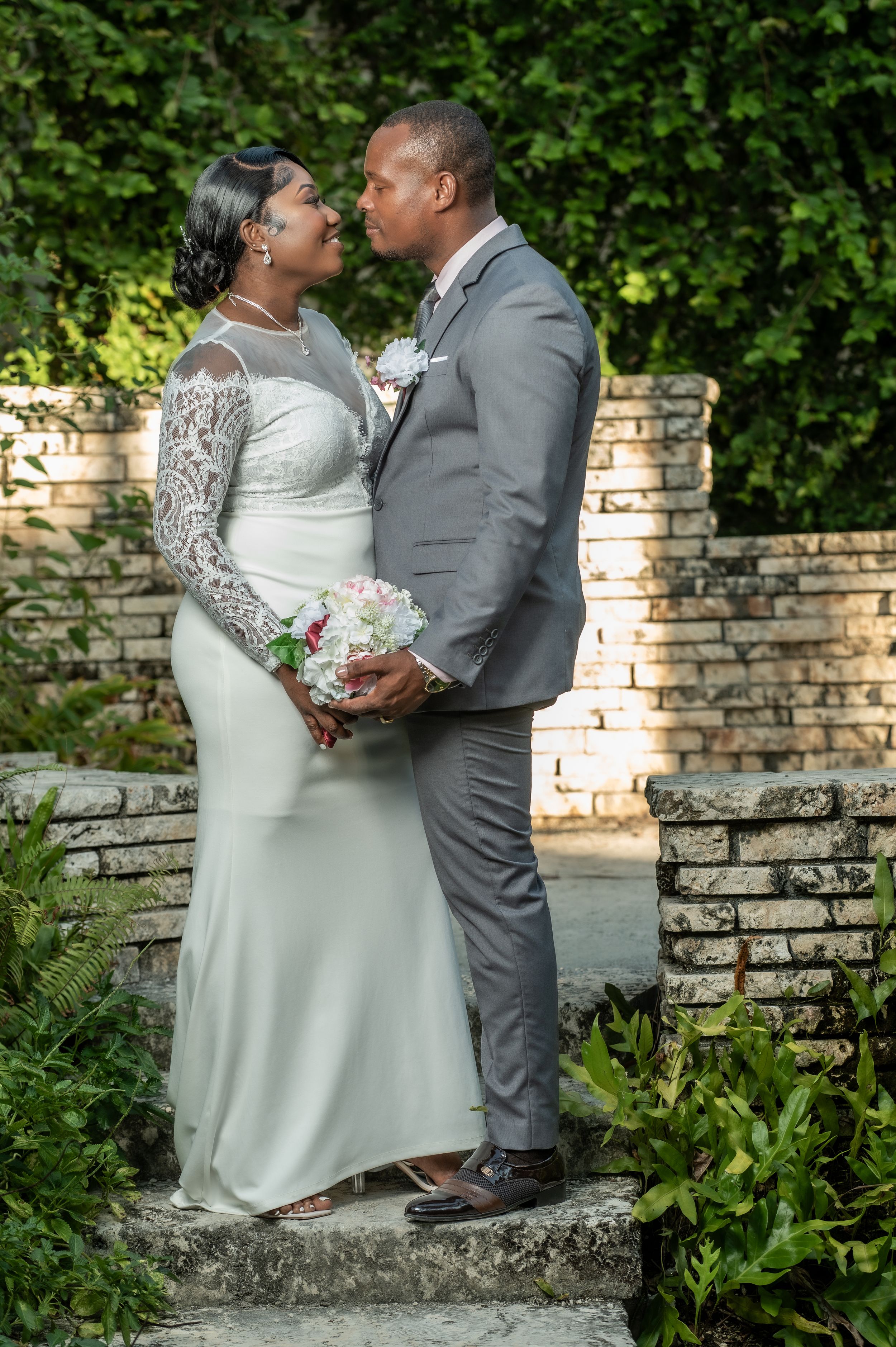 A bride and groom share a romantic kiss in a lush garden setting, she in a white gown, he in a gray suit.