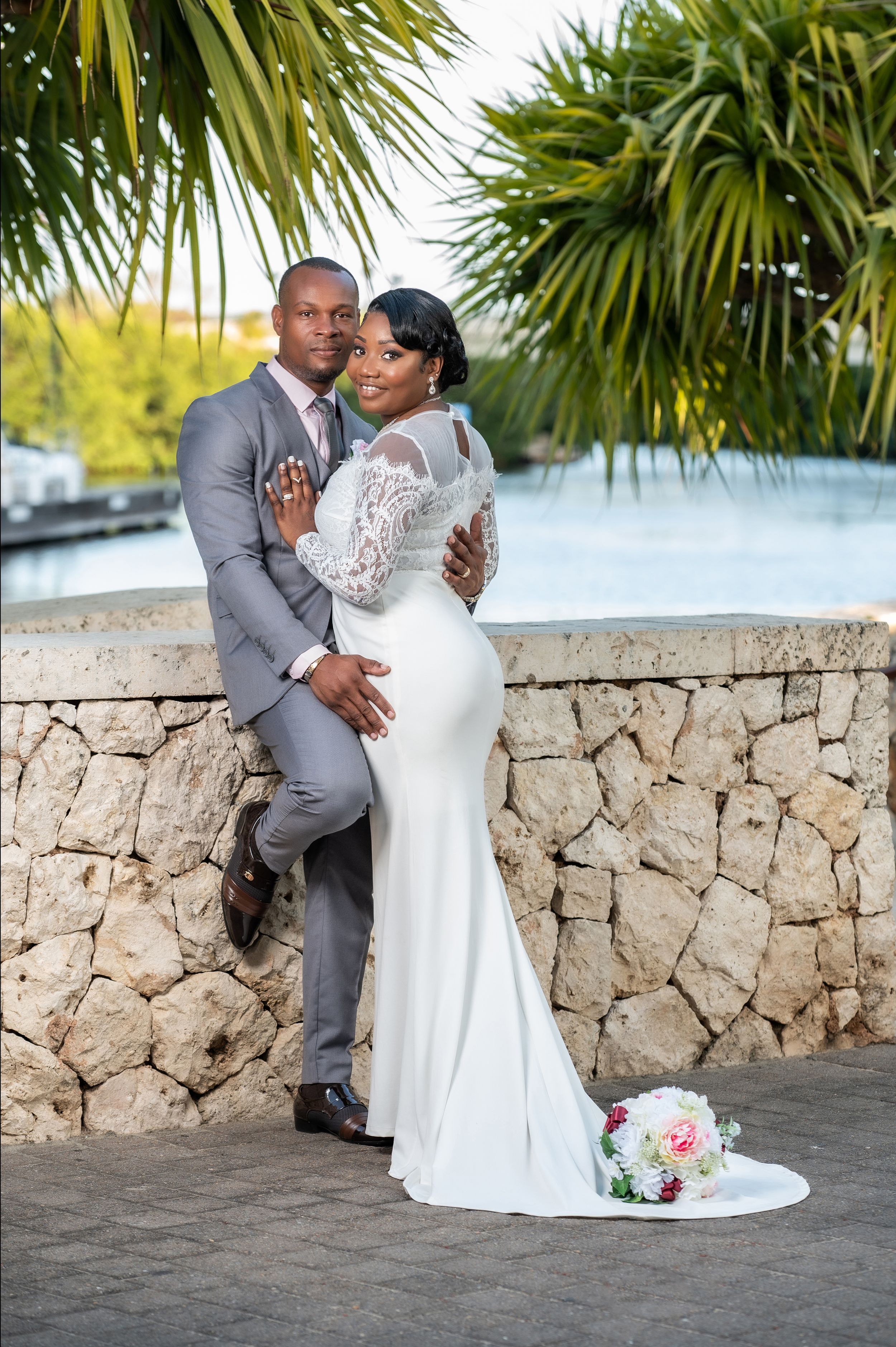 Elegant bride and groom posing outdoors by a stone wall with tropical palms during their destination wedding.
