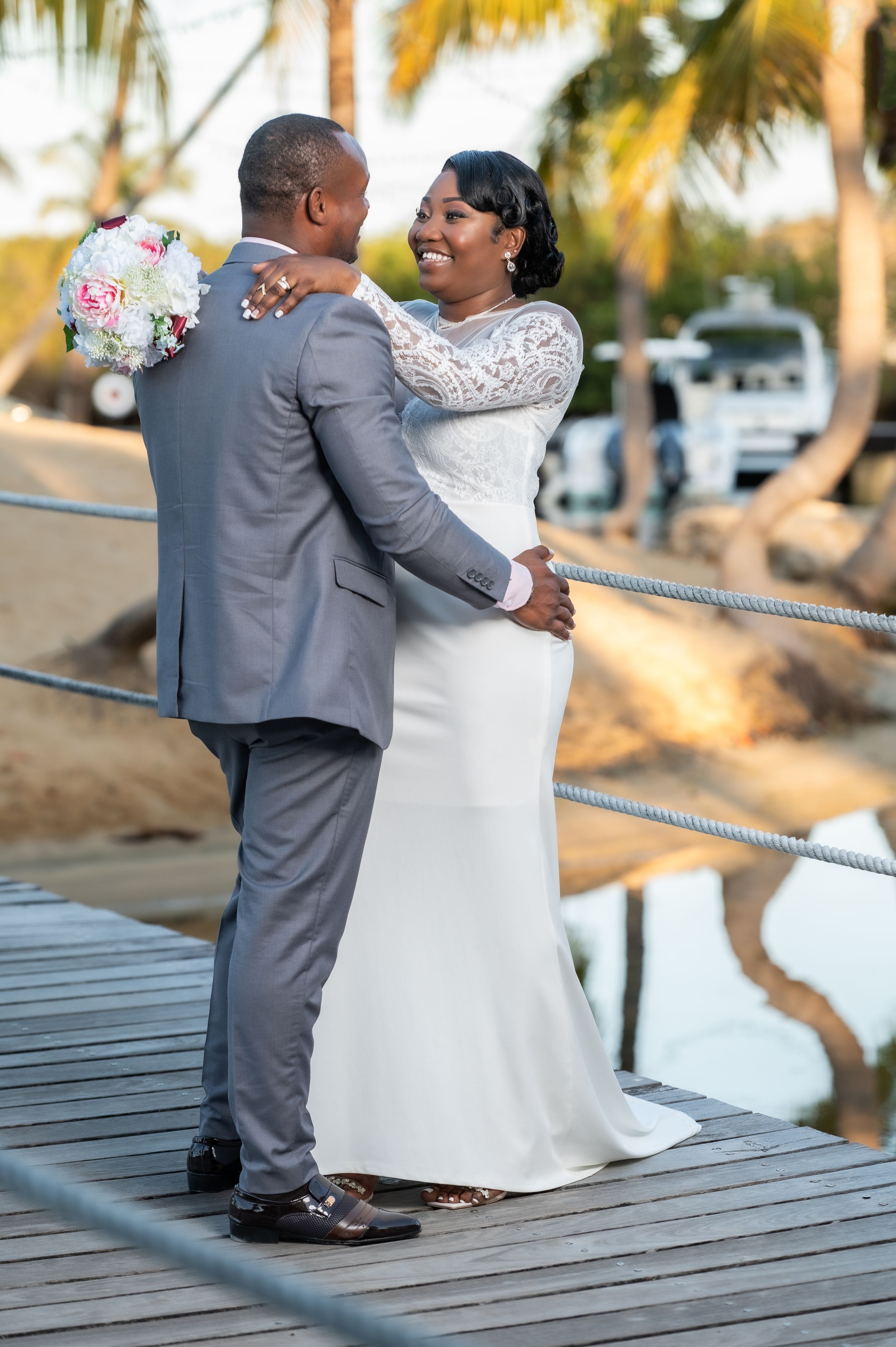 A bride and groom share a romantic moment on a wooden dock, she in a lace gown holding a bouquet, he in a gray suit.