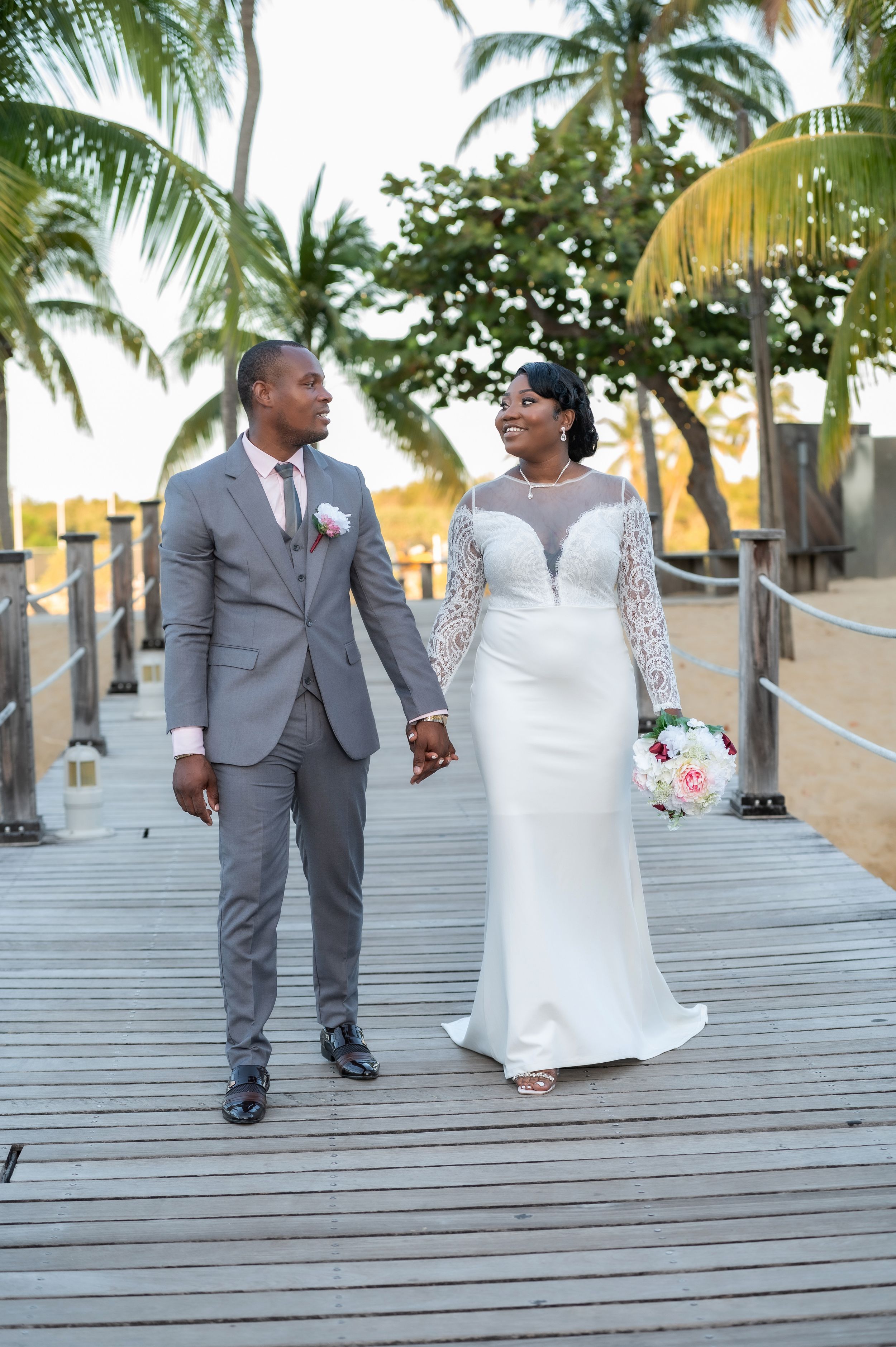 Newlywed couple in wedding attire holding hands on a wooden boardwalk surrounded by palm trees at sunset.