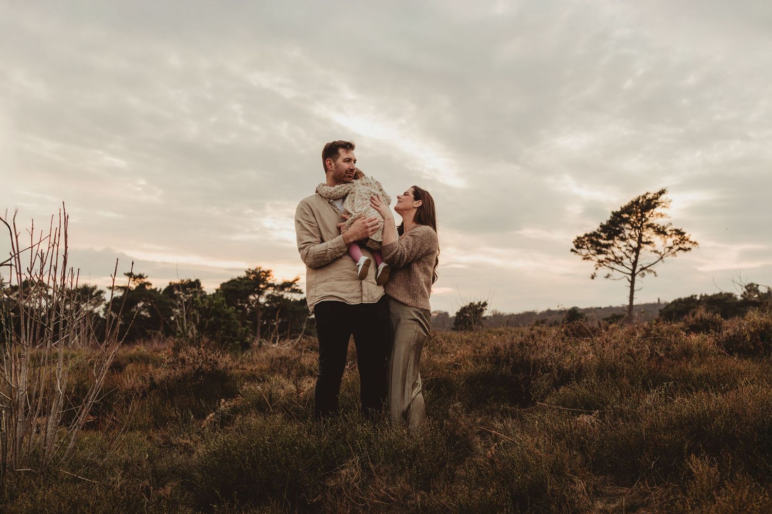 A couple embraces on a hilltop during golden hour with a lone pine tree and moody sky in the background.