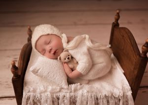 Sleeping newborn in white knit outfit on antique wooden doll bed.