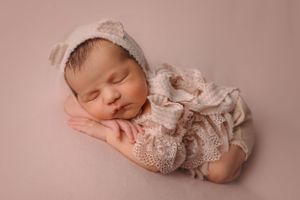 A sleeping newborn baby wearing a lace outfit and white headband rests peacefully on a pink backdrop.