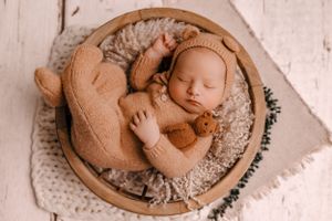 Sleeping newborn in tan knitted outfit nestled in wooden bowl filled with soft gray blanket.