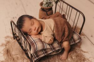 Newborn sleeping on plaid cushions in a small vintage metal bed frame with brown textured fabric.