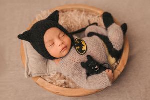 Sleeping newborn dressed in Batman-themed costume lying in wooden bowl.